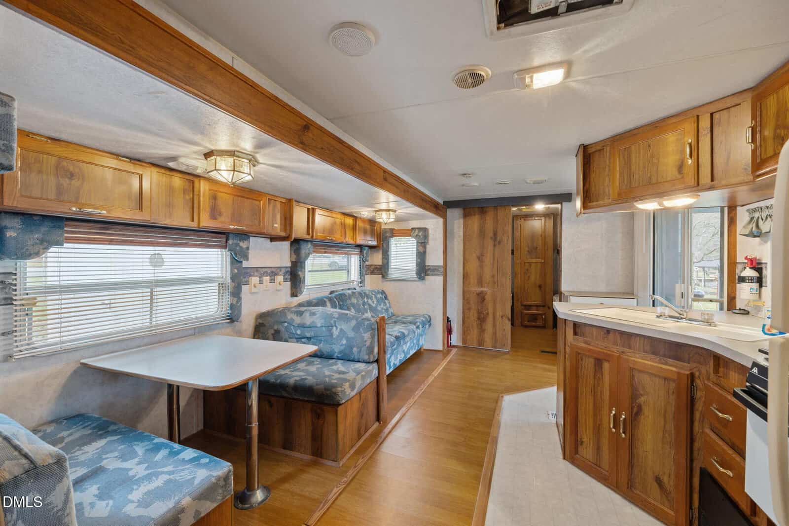 The interior of an RV with wood paneling, blue patterned bench seating around a table, wood cabinets, a small kitchen area with sink and stove, windows with blinds, and a door at the end of the space.