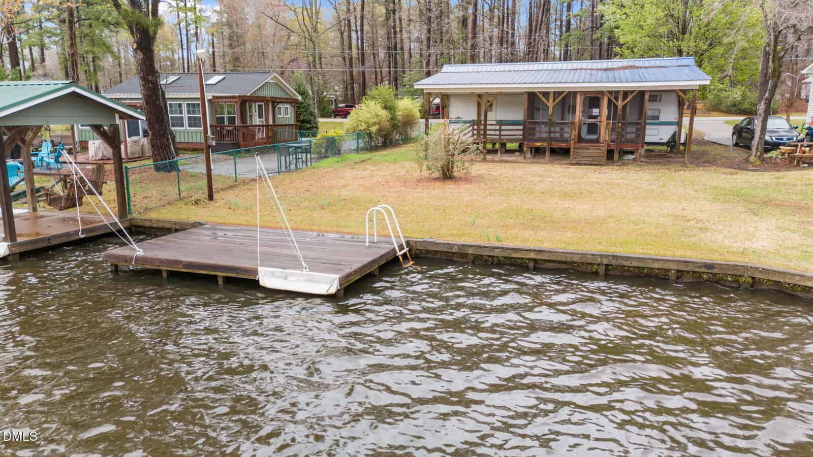 A small wooden cabin with a porch is situated on a grassy lot by the water, with a wooden dock and a small boat moored beside it; other cabins and trees are visible in the background.