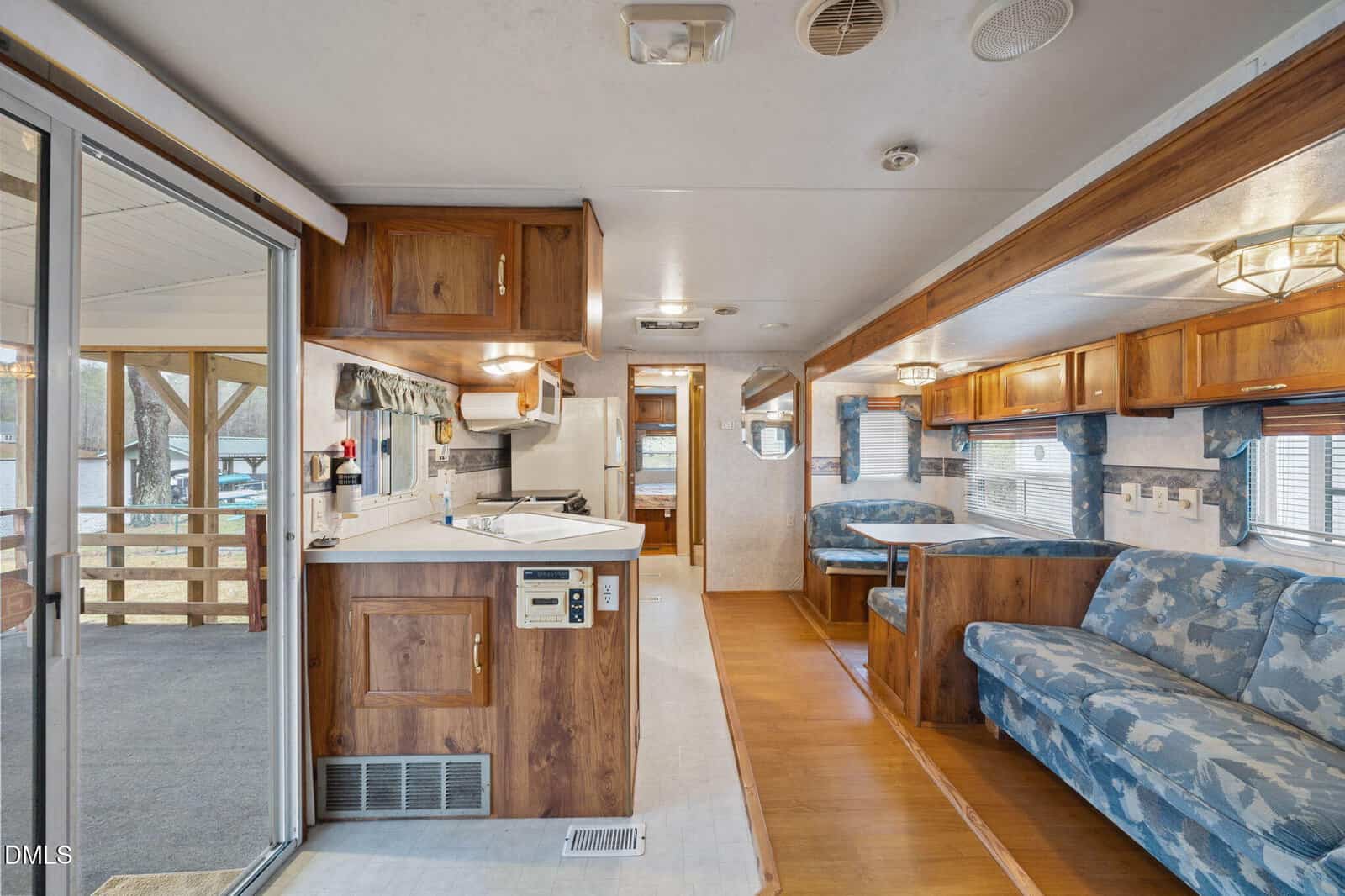 The interior of an RV with wood cabinets, a small kitchen, blue patterned couches, and a dining area. Large windows line one side, and a glass sliding door opens to a covered porch outside.