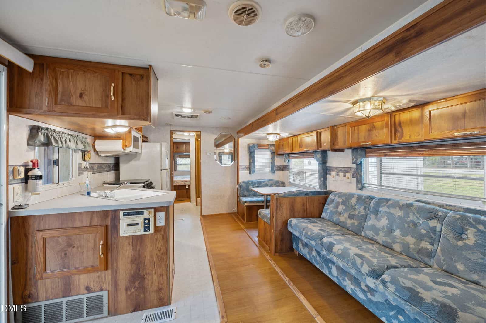 Interior of an RV with wood cabinetry, a compact kitchen on the left, blue patterned sofas on the right, a dining booth at the far end, and windows along both sides letting in natural light.