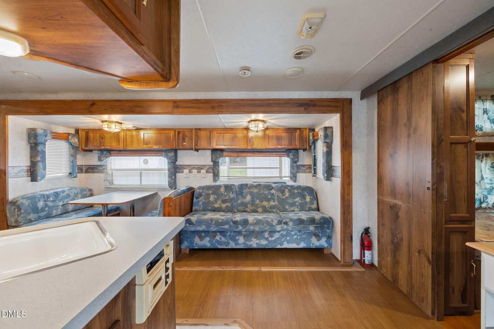 Interior of an RV showing a blue patterned sofa, a dinette with two benches, wood cabinets, and a fire extinguisher near a wooden door. Light fixtures and windows with blinds are also visible.
