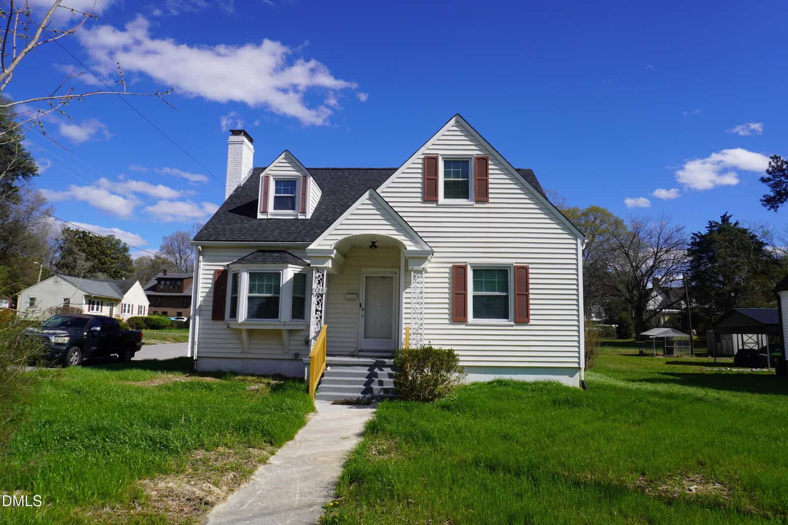 A small, two-story white house with a dark roof, red shutters, and a front porch sits on a grassy lawn under a partly cloudy sky. A concrete path leads from the sidewalk to the front steps.