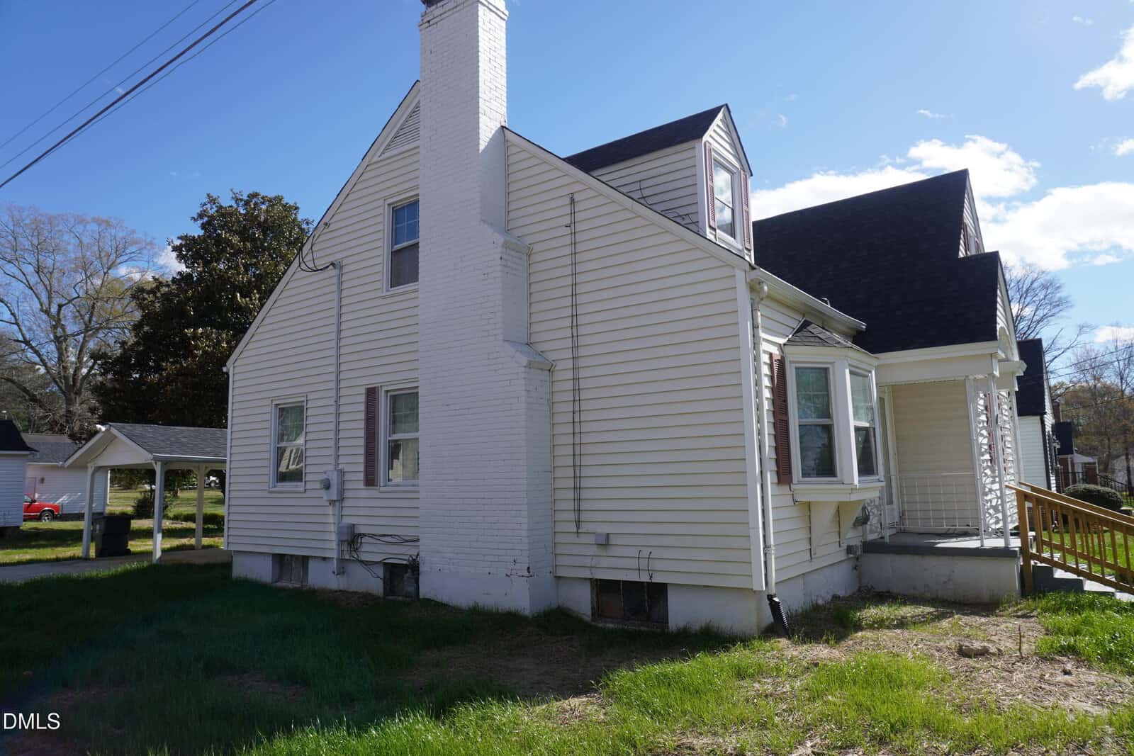 A two-story white house with vinyl siding, a prominent white brick chimney, multiple windows, and a small front porch with a wooden ramp. The yard has patchy grass, and power lines are visible overhead.