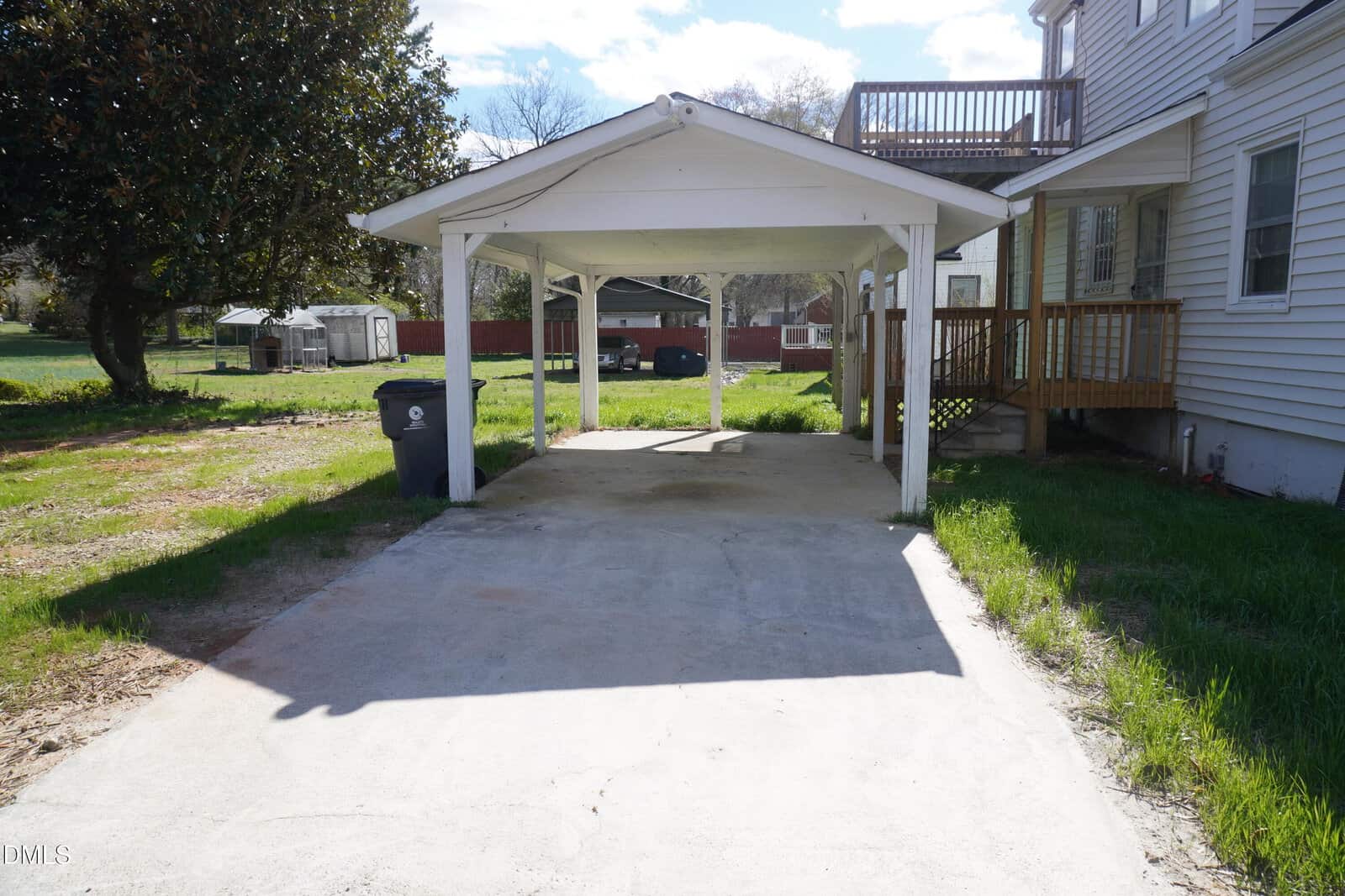A detached carport with a concrete driveway stands beside a white house with siding. A trash bin is on the left under the carport, and there is a small wooden deck attached to the house on the right.