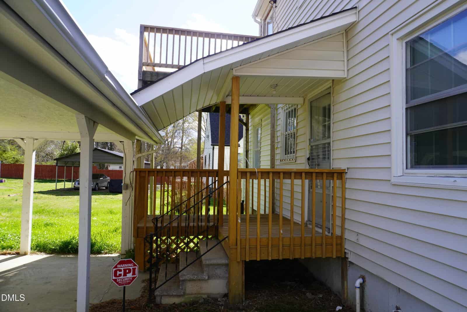 A wooden porch with black metal railings leads to the back entrance of a beige house with vinyl siding. An upper balcony is visible above. A security sign is posted in the grassy yard near a covered carport.