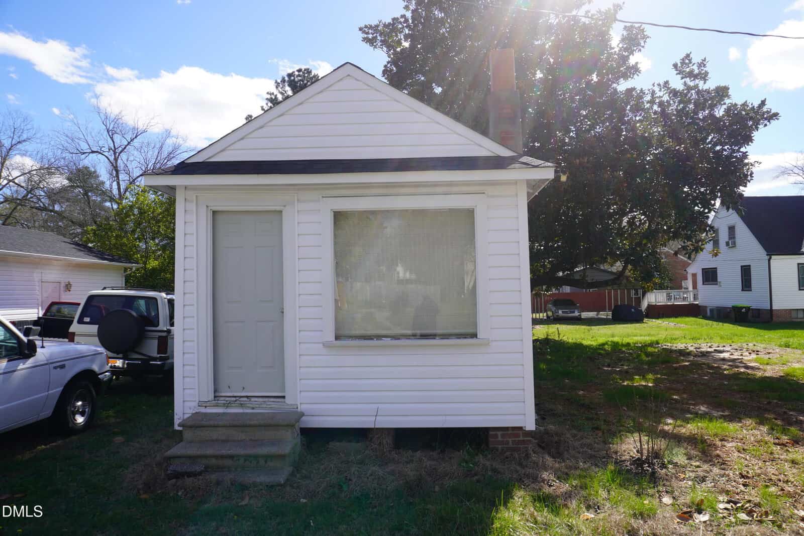 A small white single-story house with a front door, large window, and steps leading to the entrance. The house has vinyl siding and sits on a grassy yard with other homes and vehicles visible in the background.