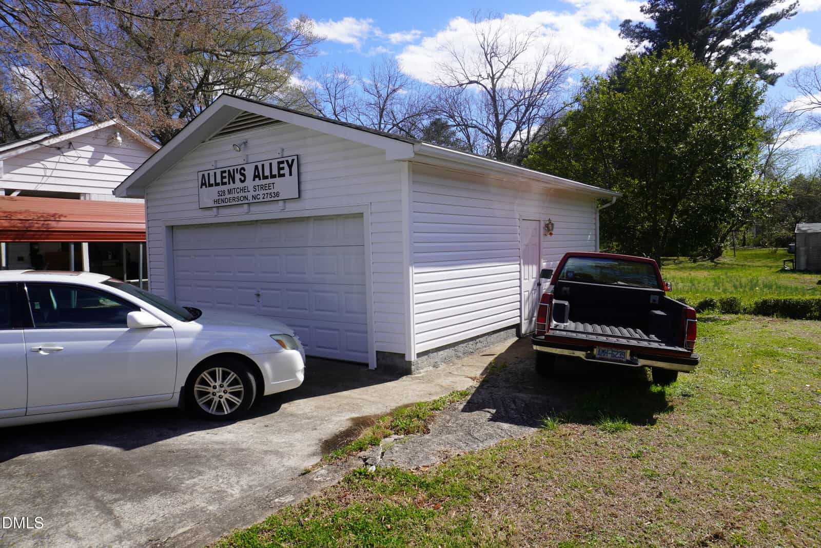 A white garage labeled “Allen’s Alley” sits beside a white house. A white car is parked in the driveway, and a black pickup truck with an open tailgate is parked on the grass nearby. Trees and grass fill the background.