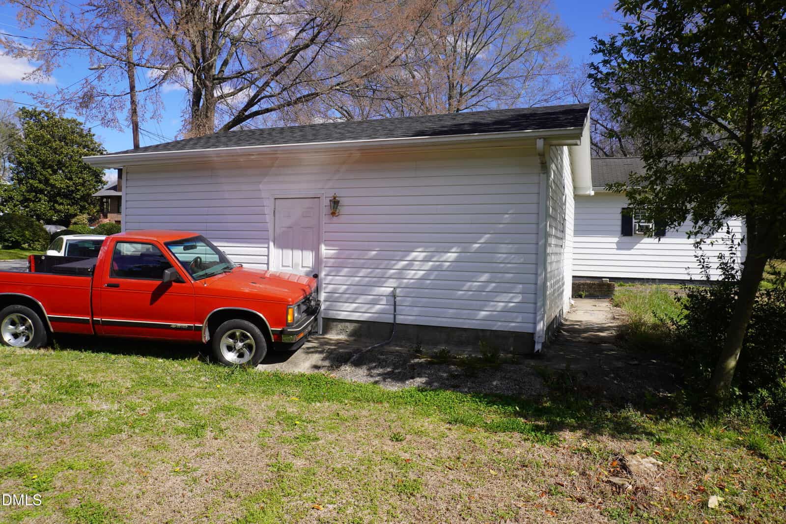 A red pickup truck is parked next to a white detached garage or shed, with trees and grass surrounding the area under a blue sky.