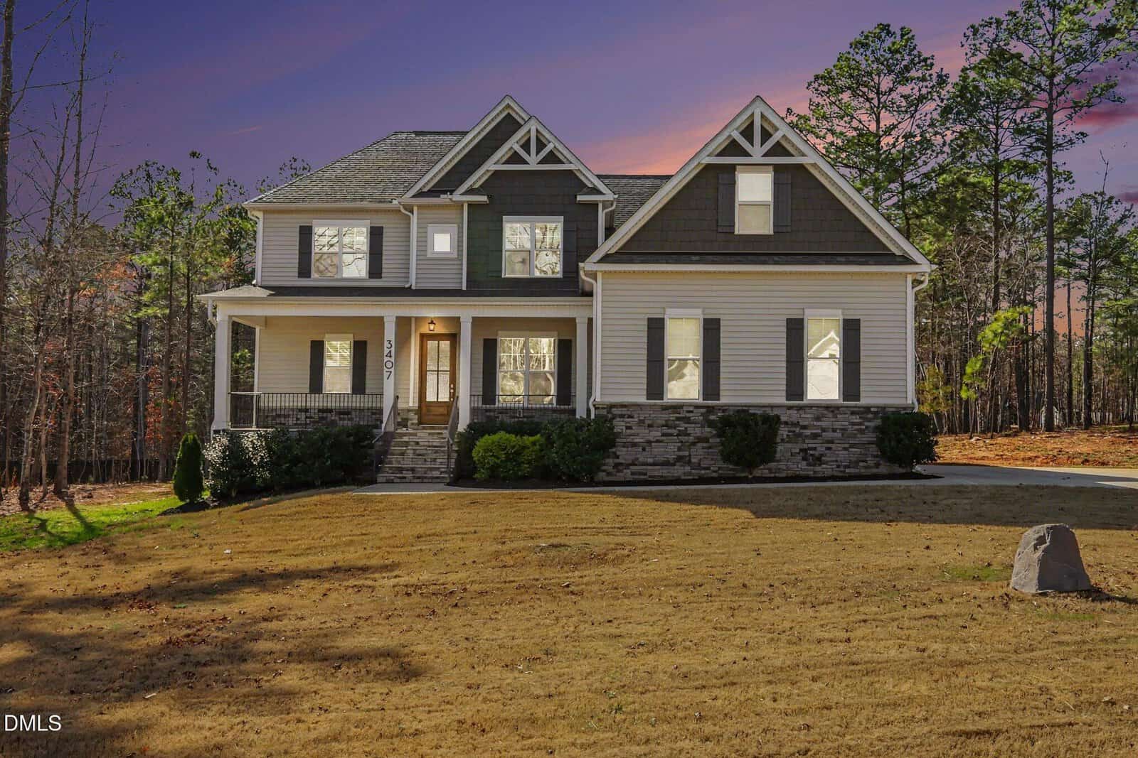 Two-story suburban house with gray siding, stone accents, and a covered front porch on a large, grassy lawn at 3407 River Manor Court, Franklinton. Surrounded by tall pine trees, the sky is colorful at sunset.