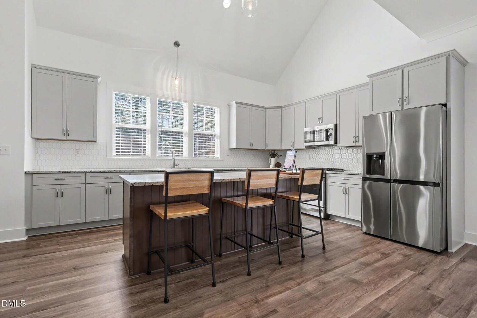 Modern kitchen with light gray cabinets, white tile backsplash, stainless steel appliances, a large island with a dark wood base, three wooden bar stools, and wood-style flooring. Sunlight streams through three windows.