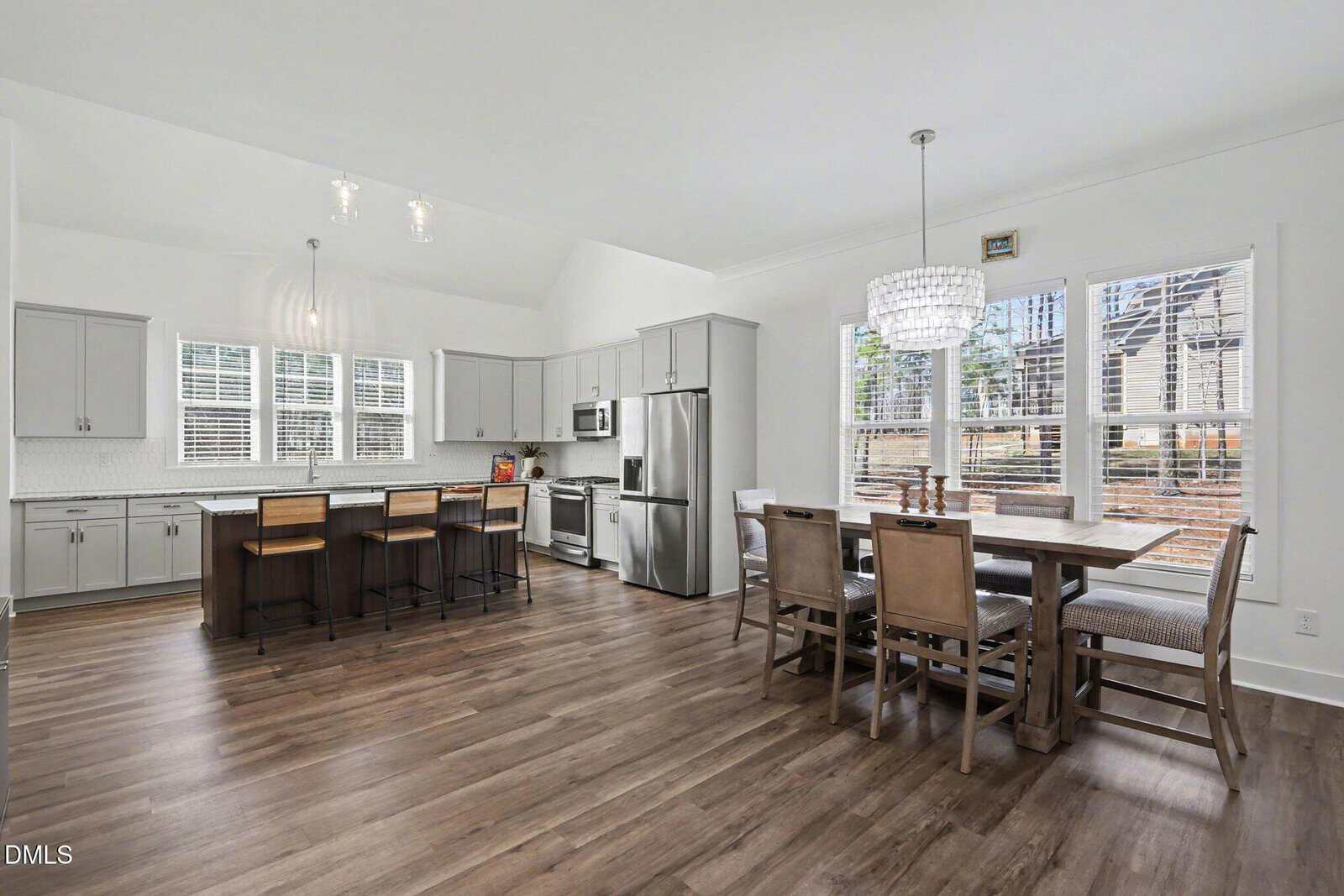 Spacious modern kitchen with gray cabinets, stainless steel appliances, a large island with barstools, and a dining table for six near large windows overlooking a wooded backyard.