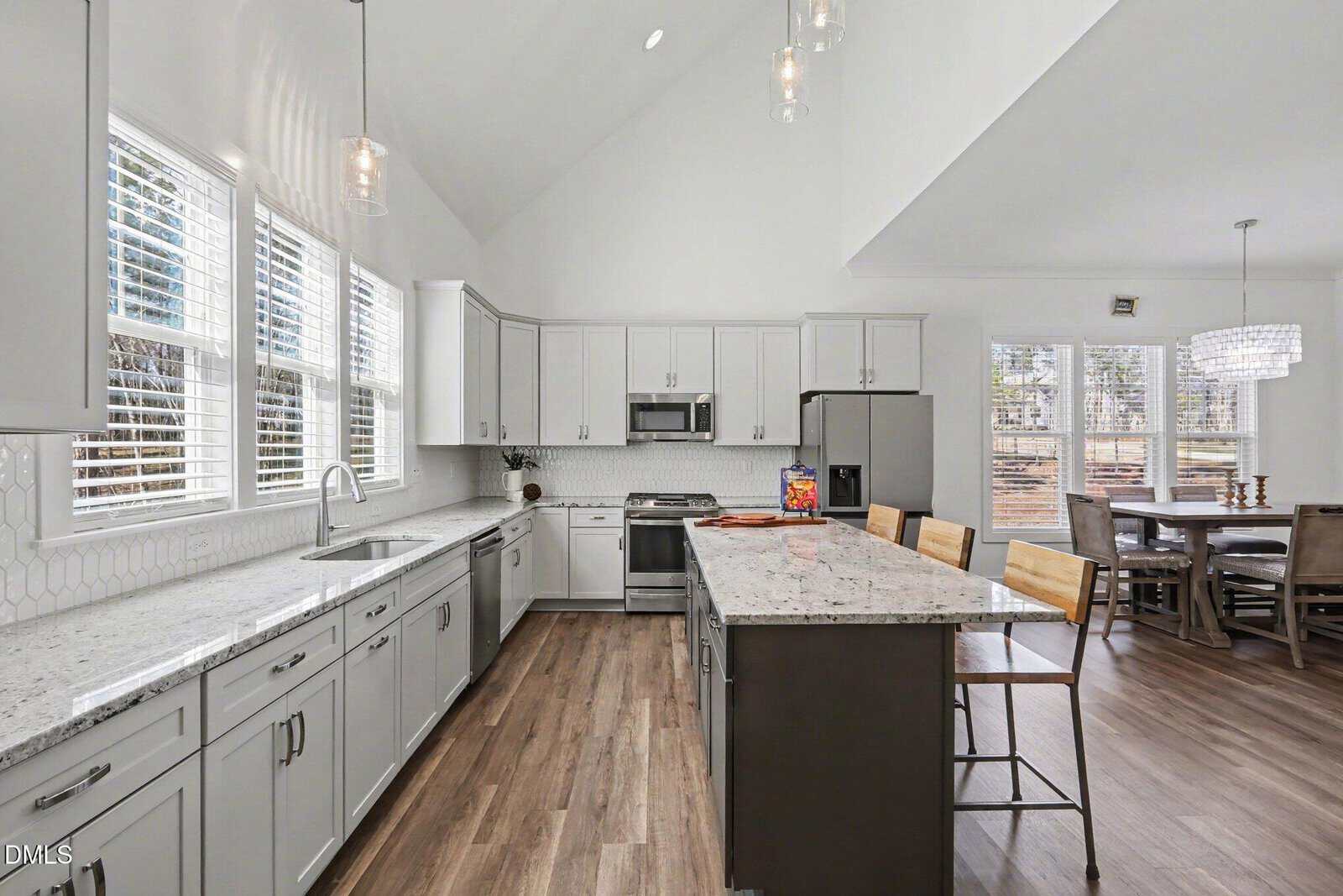 Modern kitchen with white cabinets, stainless steel appliances, a large island with four wooden barstools, pendant lighting, and a dining area with a chandelier and windows overlooking trees in the background.