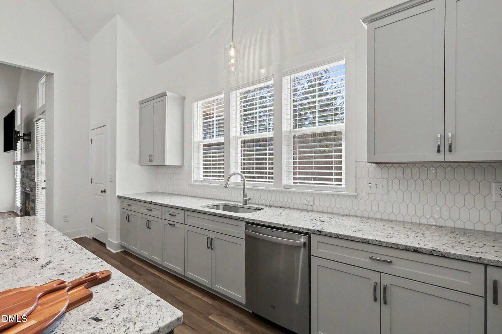 Modern kitchen with light gray cabinets, white granite countertops, stainless steel dishwasher, and large windows with white blinds. White hexagonal tile backsplash and a pendant light above the sink.