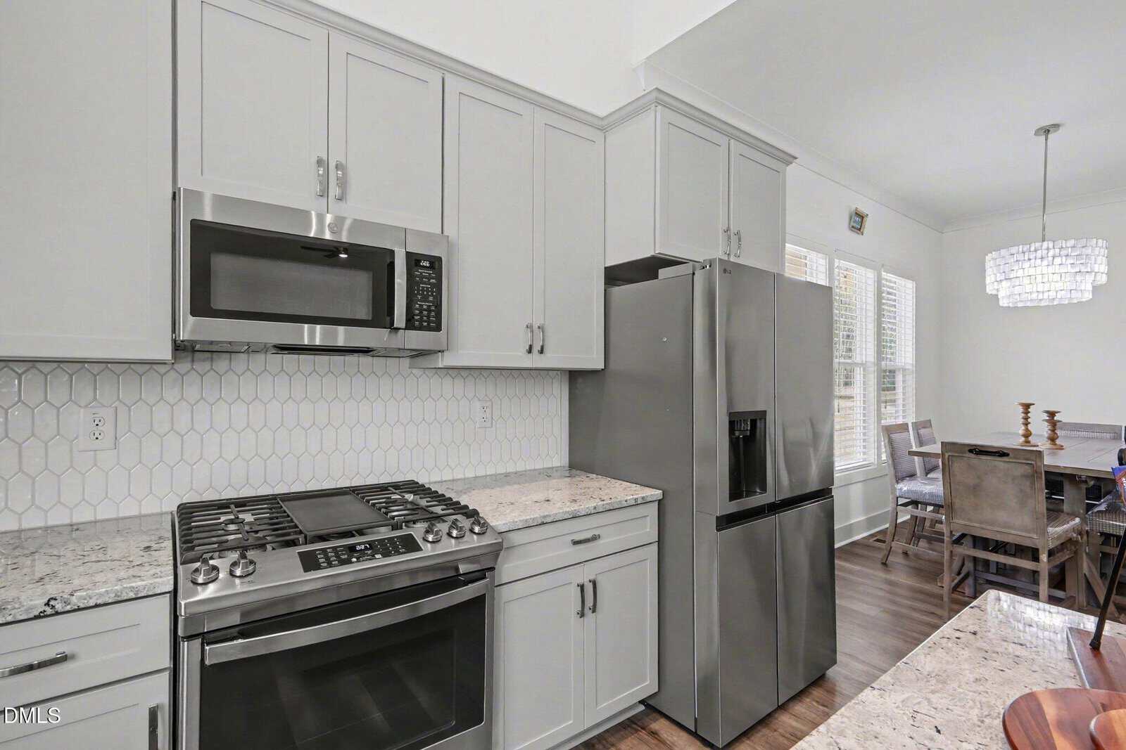 Modern kitchen with gray cabinets, stainless steel appliances including a gas stove, microwave, and refrigerator, white hexagonal tile backsplash, and a dining area visible in the background.