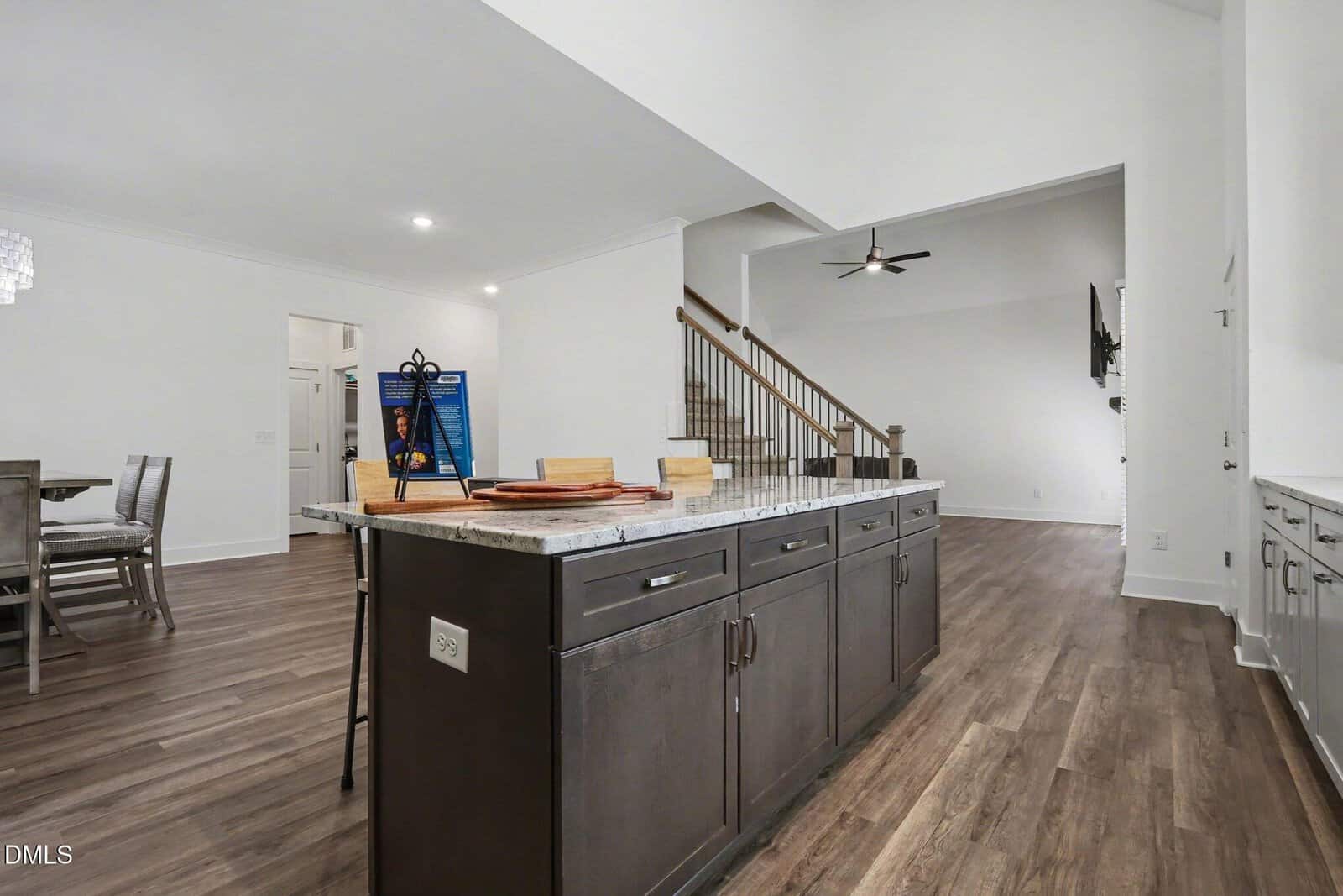 A modern kitchen with dark cabinets and a granite island countertop, overlooking an open dining area and living room with wood flooring and a wooden staircase in the background.