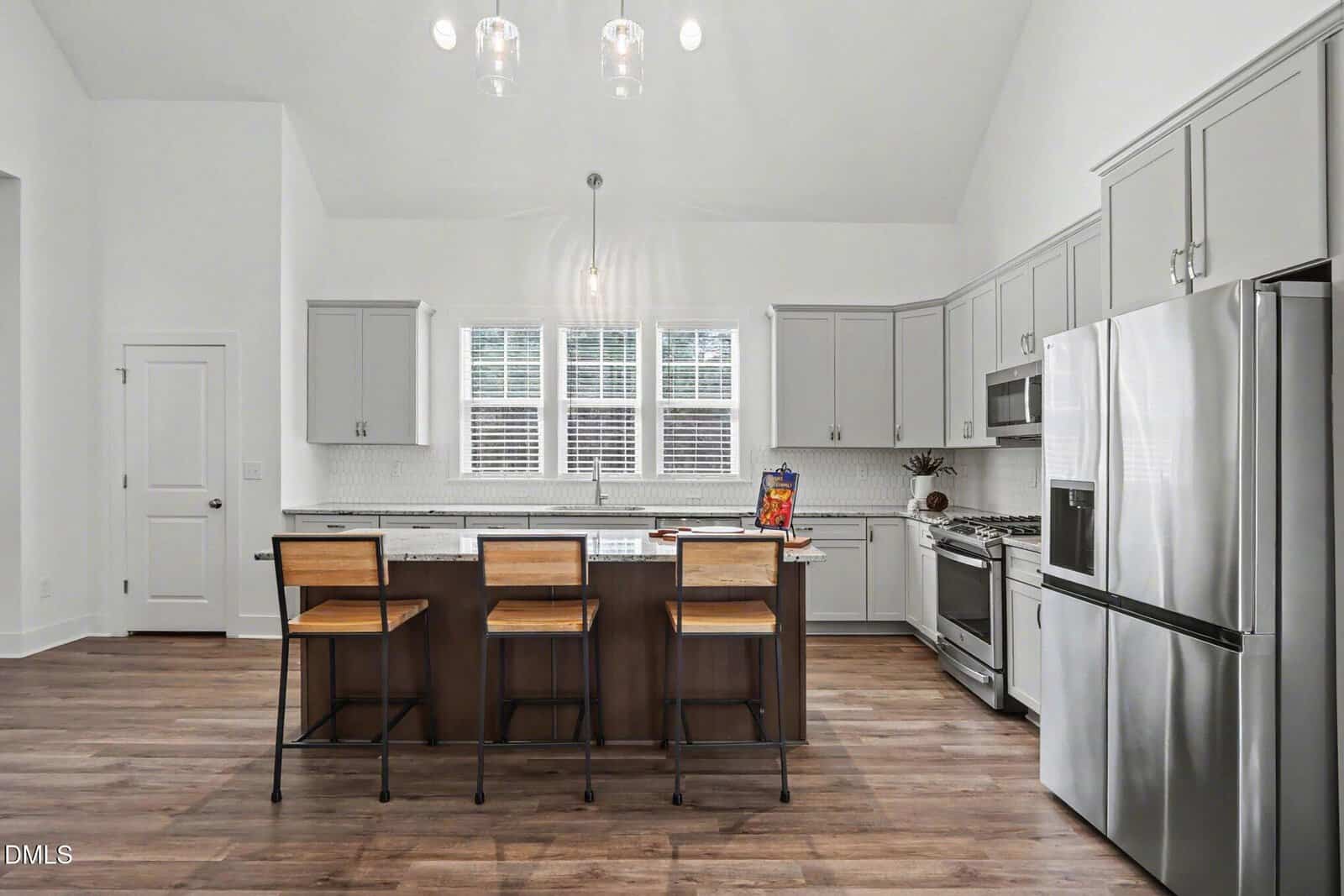 Modern kitchen with gray cabinets, stainless steel appliances, a large island with three wooden barstools, pendant lights, and three windows above a sink. A cereal box and bowl are on the island countertop.