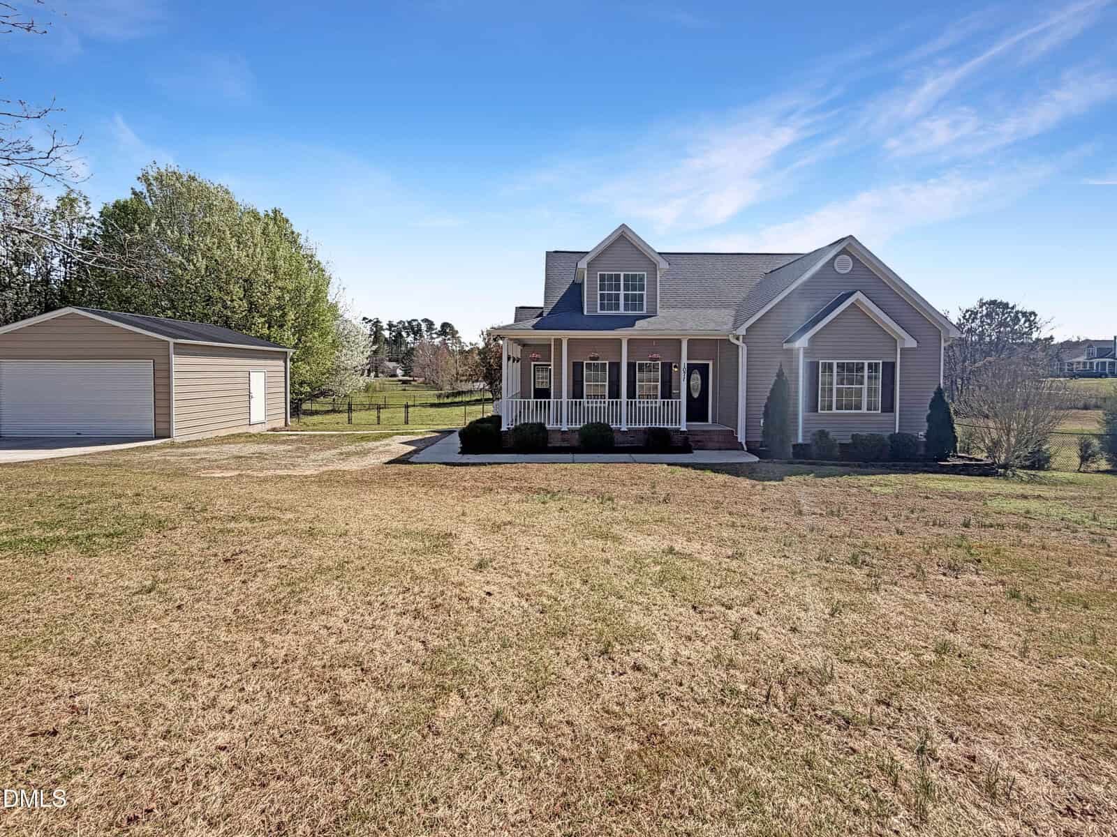 A single-story house with a covered front porch and dormer windows sits on a large grassy lot with a detached garage to the left. The sky is mostly clear with a few clouds.
