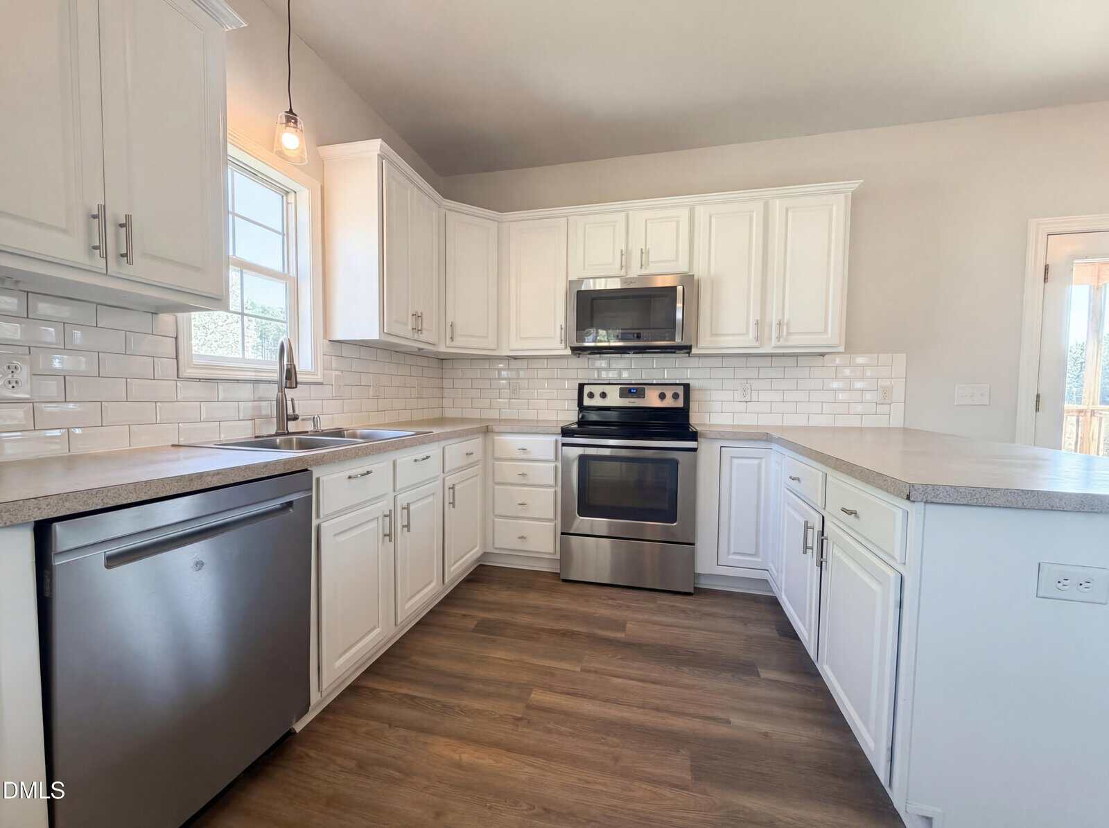 Modern kitchen with white cabinets, stainless steel appliances, subway tile backsplash, light countertops, a window over the sink, wood-look flooring, and a pendant light above the sink area. Door to the right leads outside.