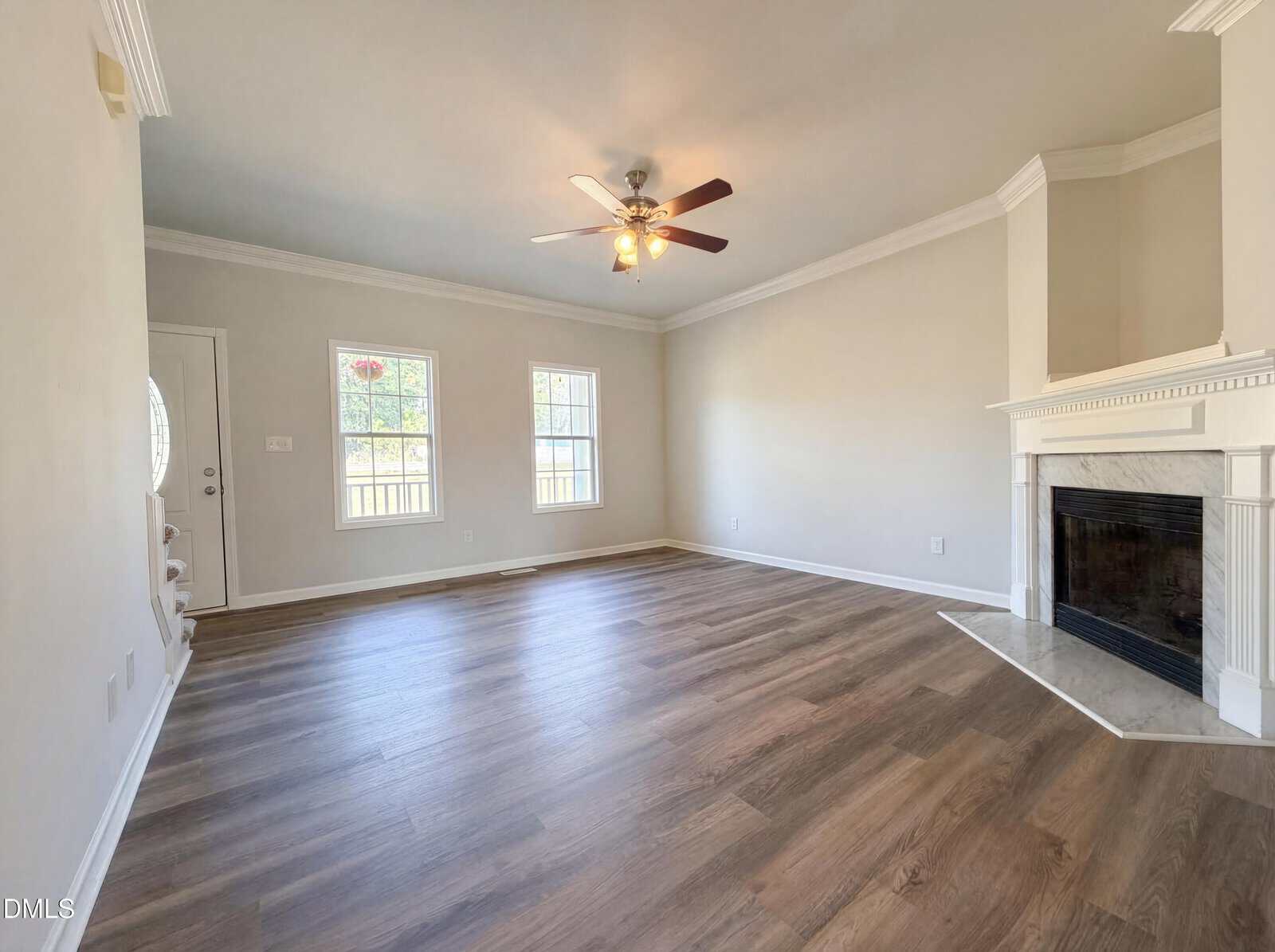 A bright, empty living room with light-colored walls, a ceiling fan with lights, two windows, wood-style flooring, and a white fireplace with a mantel in the corner.