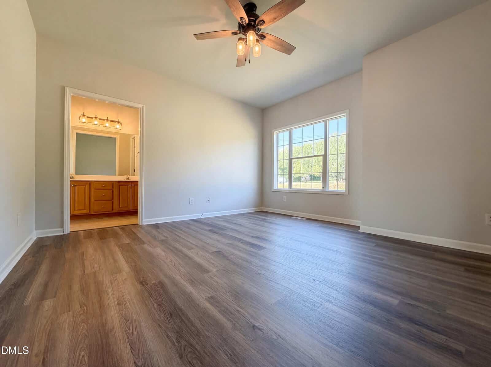 Empty bedroom with wood flooring, a ceiling fan with lights, a large window, and an open doorway leading to a bathroom with a double-sink vanity and mirror. Walls are light-colored and the room is well-lit by natural light.