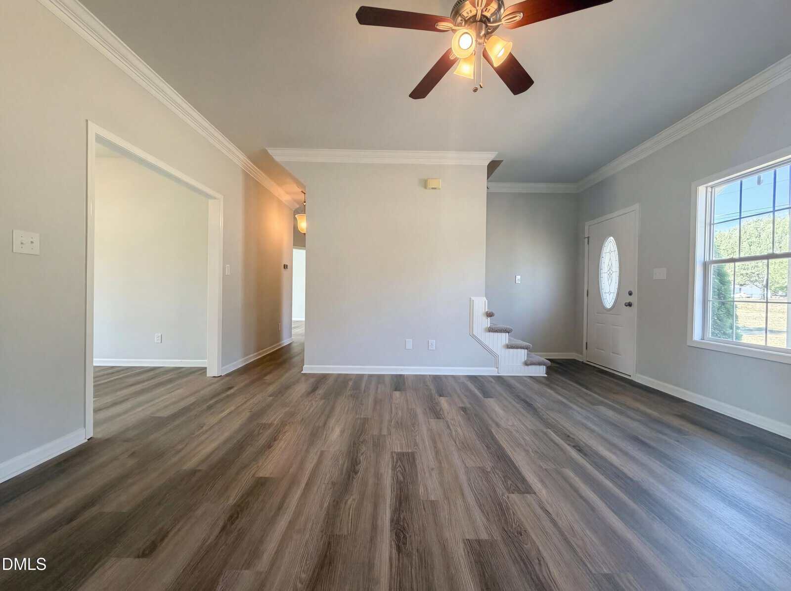 A spacious, empty living room with light gray walls, wood-look flooring, a ceiling fan with lights, white trim, and a window and door on the right side; a staircase leads upward near the door.