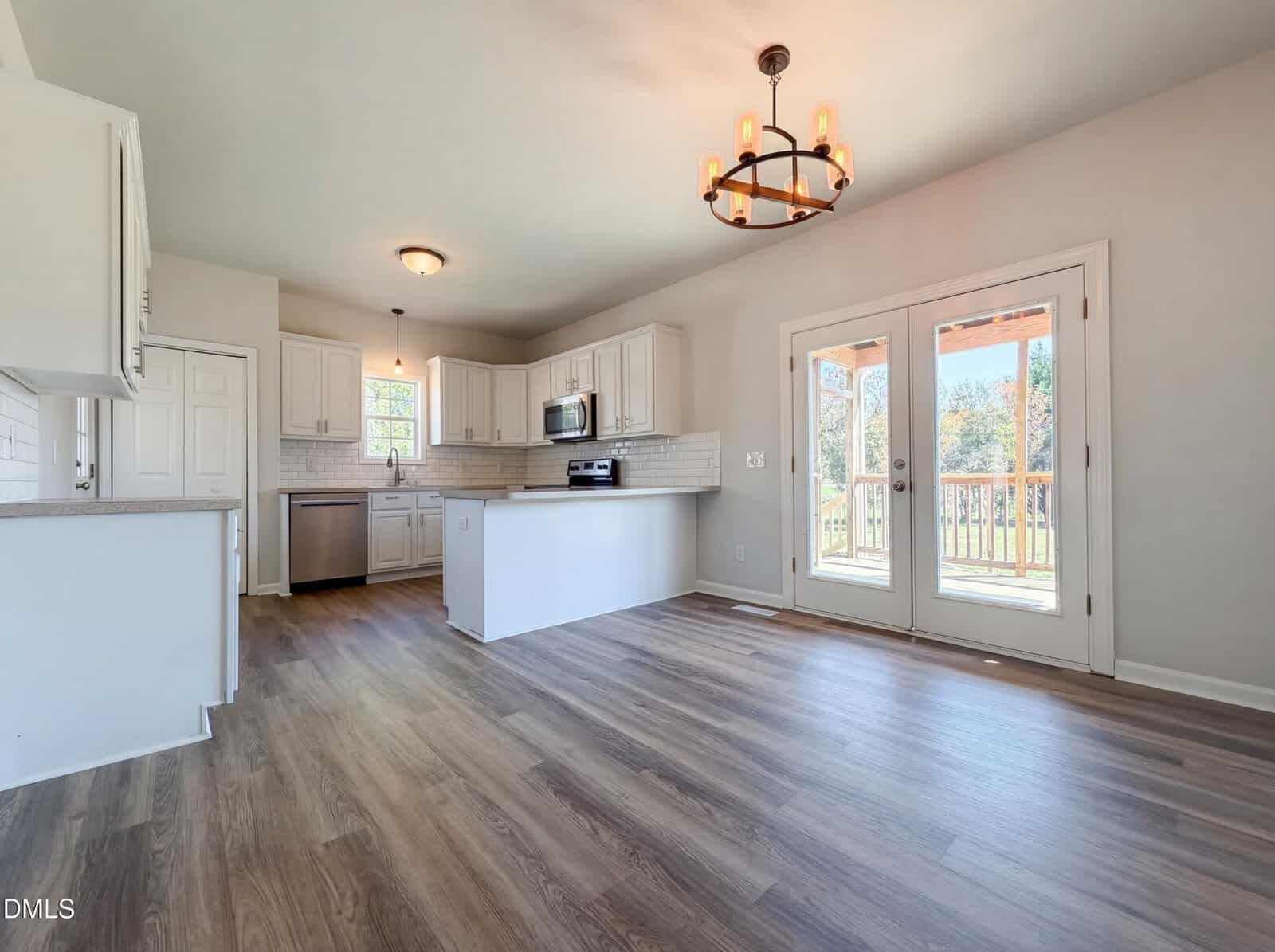 Bright, modern kitchen and dining area with white cabinets, stainless steel appliances, wood-style flooring, a black chandelier, and double glass doors leading to a deck with a view of trees outside.