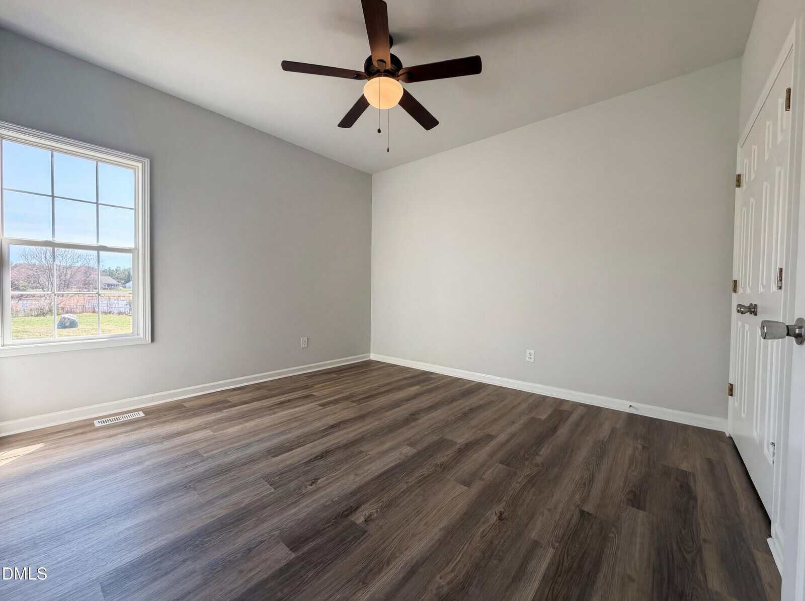 Empty room with light gray walls, wood-style flooring, a ceiling fan with a light, a window showing an outdoor view, and double white doors. Sunlight enters through the window, brightening the space.