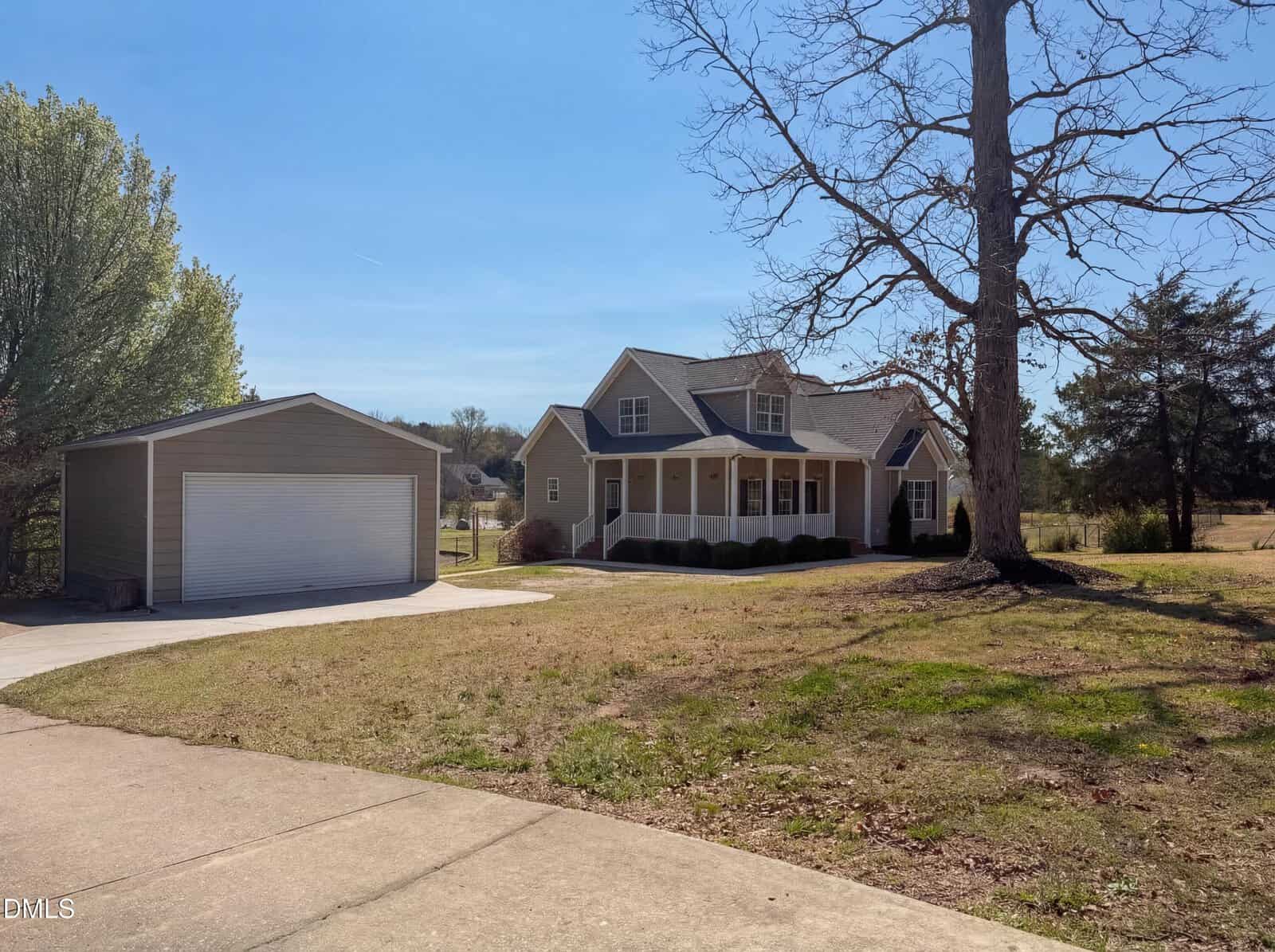 A beige house with a covered front porch and dormer windows sits beside a detached single-car garage. The property has a lawn with a large tree, a concrete driveway, and is bordered by trees.