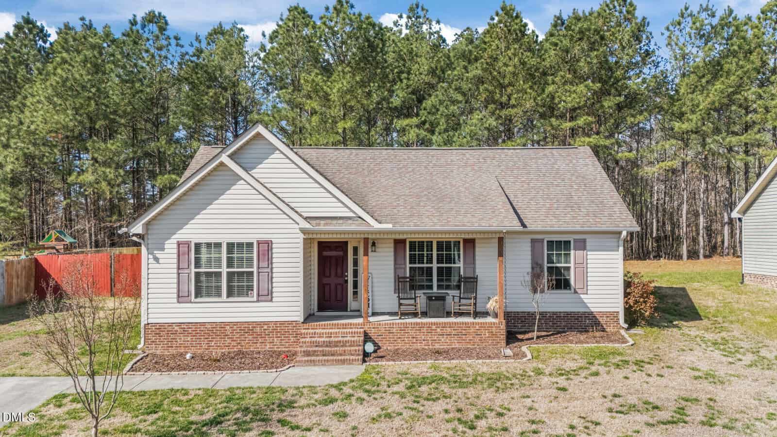 A single-story house at 204 Orkney Road in Stem features beige siding, a brick foundation, and a covered front porch with chairs, set against tall pine trees and a partially fenced yard.