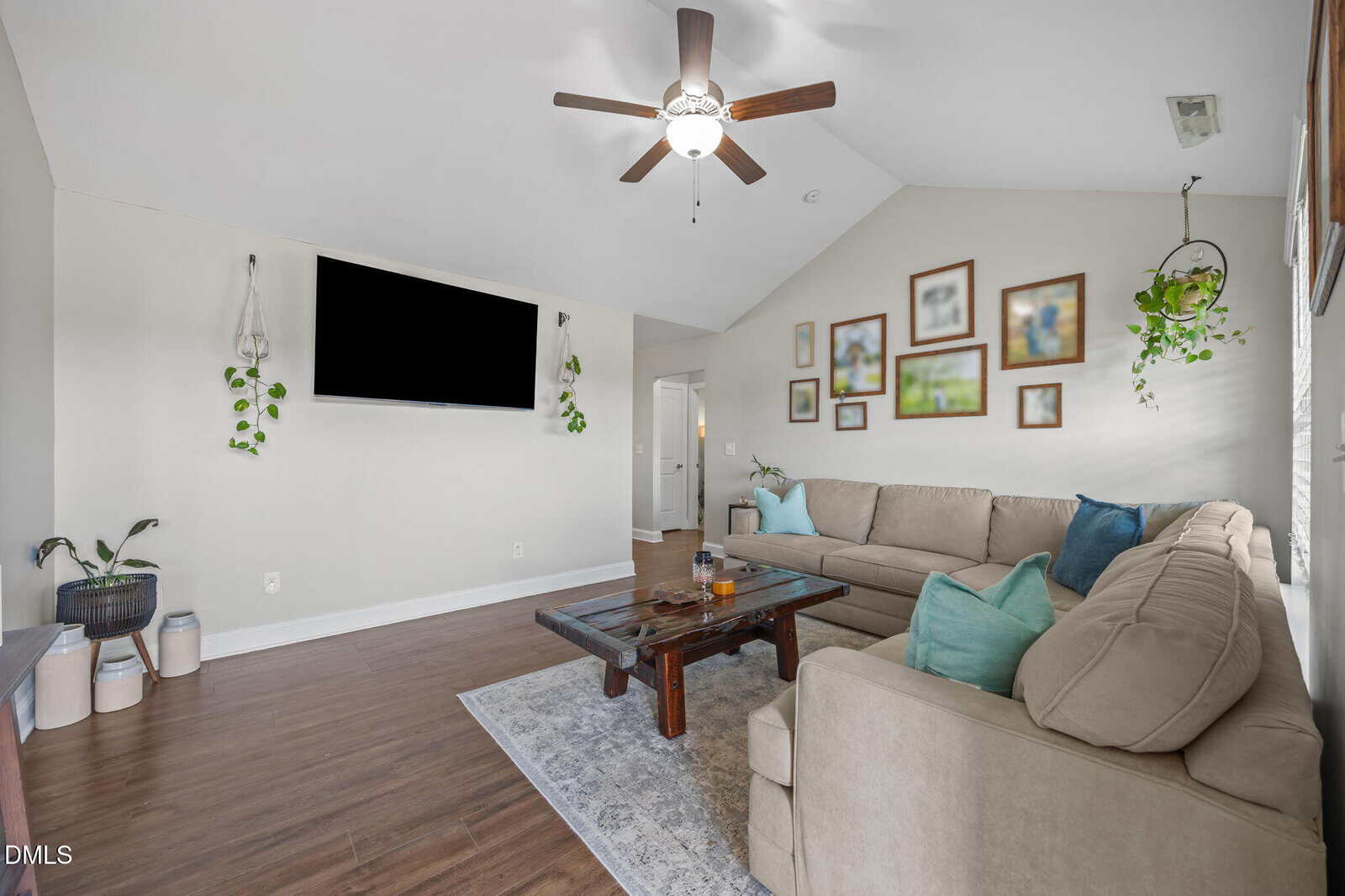 A bright living room with a beige sectional sofa, blue and green pillows, a wooden coffee table, wall-mounted TV, hanging plants, wooden floors, and framed art on a light-colored wall under a ceiling fan.