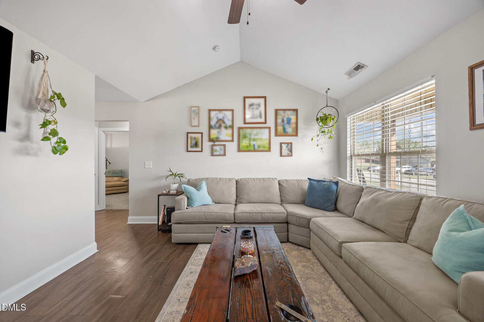 A bright living room with a large beige sectional sofa, blue and white pillows, wooden coffee table, wall art, hanging plants, and large windows letting in natural light. A TV is mounted on the left wall.