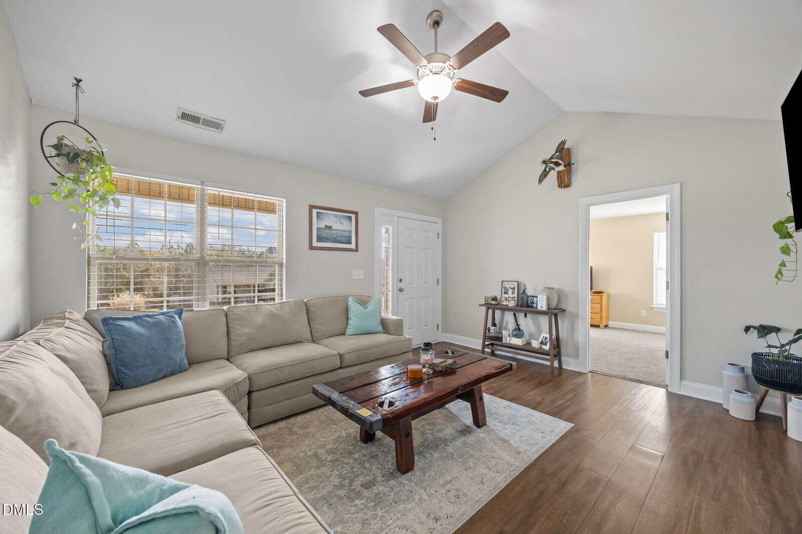 A bright living room with a beige sectional sofa, blue and white pillows, a wooden coffee table, a ceiling fan, wood-look floors, indoor plants, and sunlight streaming in through large windows.