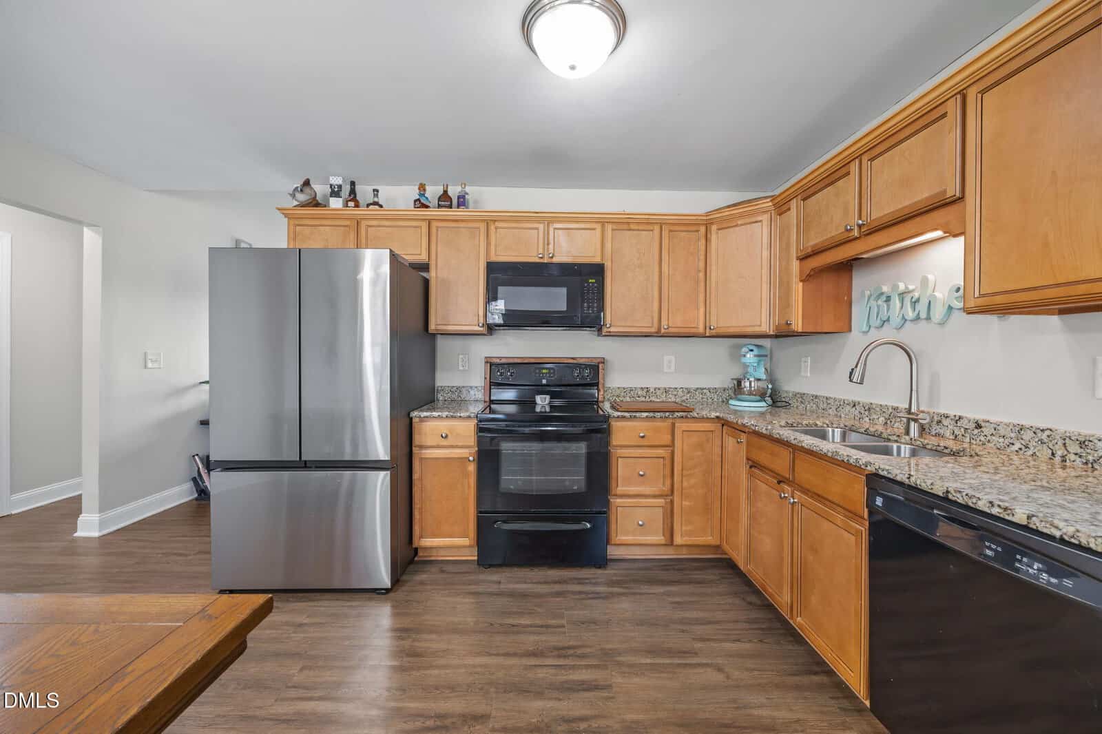 Modern kitchen with stainless steel refrigerator, black stove and microwave, wooden cabinets, granite countertops, dishwasher, and wood flooring. Small kitchen appliances and decor items are on the counters and cabinets.