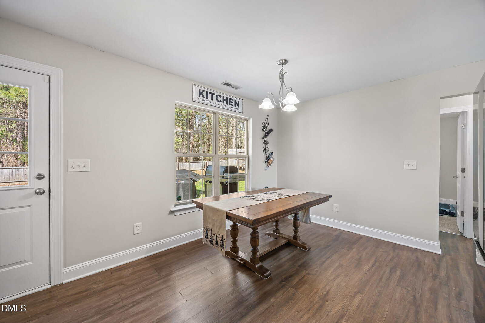 A dining area with a wooden table, a light fixture above, a window with a “KITCHEN” sign, and a door to the left. The room has light-colored walls and wood flooring. There’s a view of a fenced yard outside.