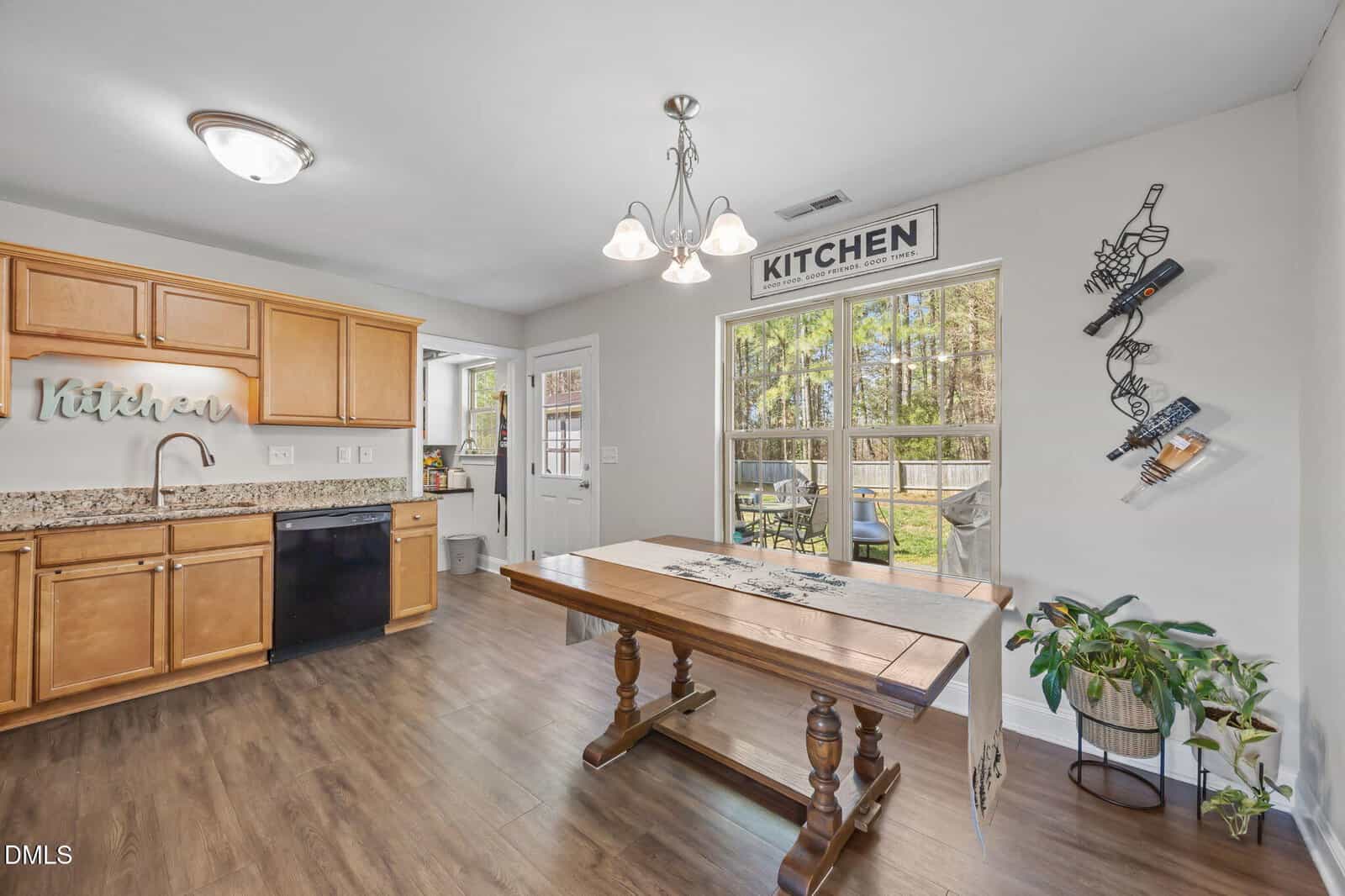 Kitchen and dining area with wood cabinets, black dishwasher, and a wooden table with a runner. Wall art spells “kitchen,” and a rack holds wine bottles. A window and door overlook a fenced backyard with trees.