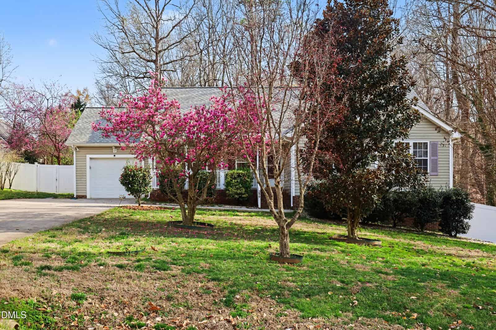 A single-story house with beige siding and a gray roof is shown behind blooming pink trees and green grass. A white garage door is visible on the left, and a white fence runs along the side and back of the property.