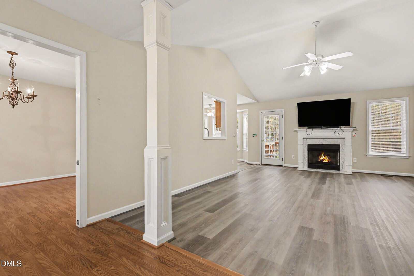 A spacious living room with light gray wood flooring, a white fireplace with a fire burning, a ceiling fan, and a wall-mounted TV. An adjacent dining area with wood flooring and a chandelier is partially visible.
