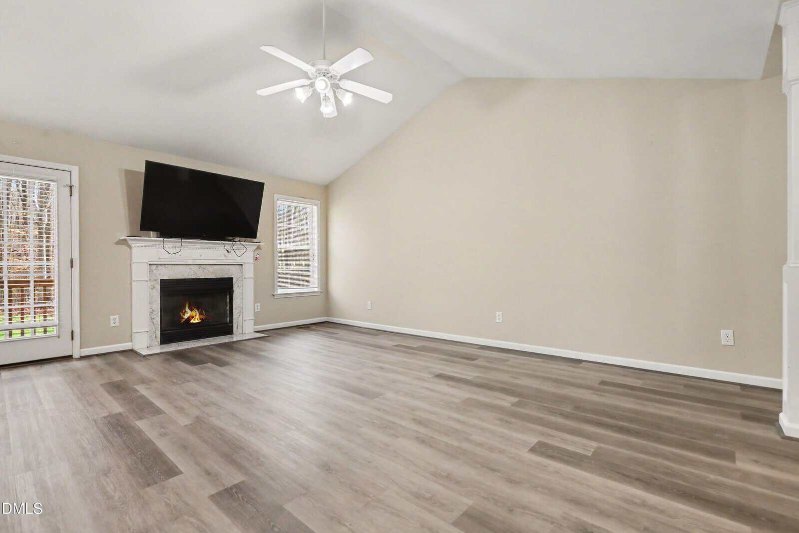 Spacious living room with light wood flooring, beige walls, a white fireplace with a fire, wall-mounted TV, ceiling fan, and windows allowing natural light. A glass door leads outside. Room is unfurnished.