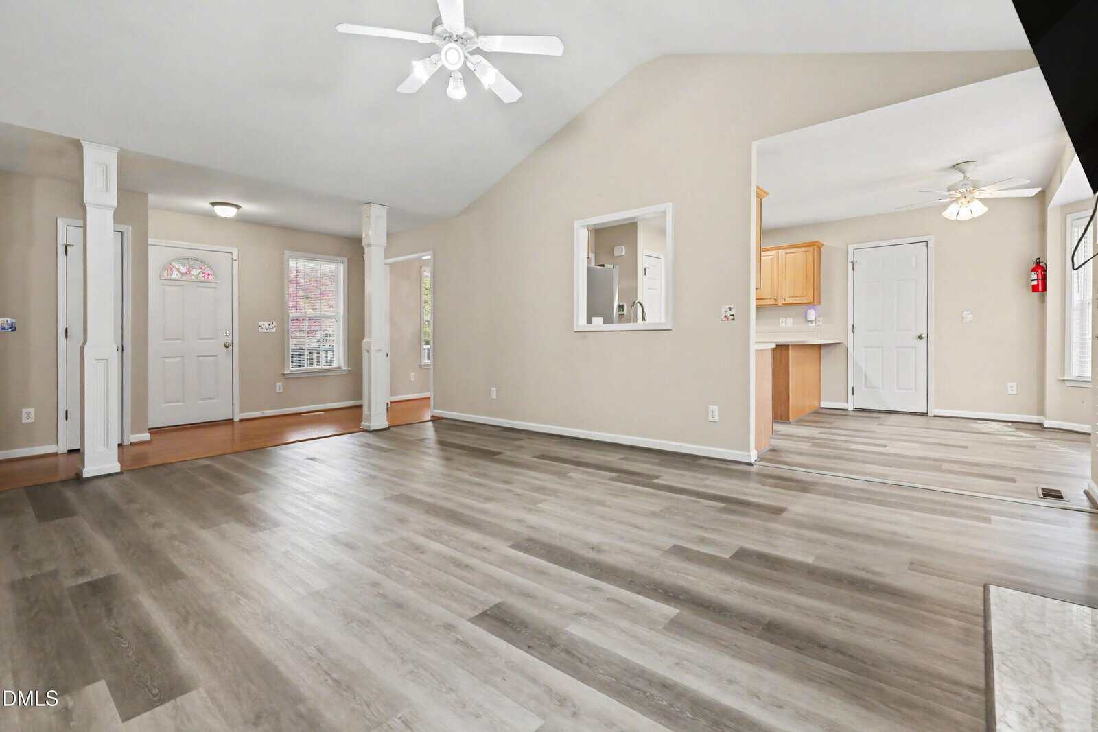 Open living space with gray wood flooring, beige walls, a ceiling fan, and windows. The area connects to a foyer with white columns and a kitchen with white cabinets and dining space. Natural light fills the room.