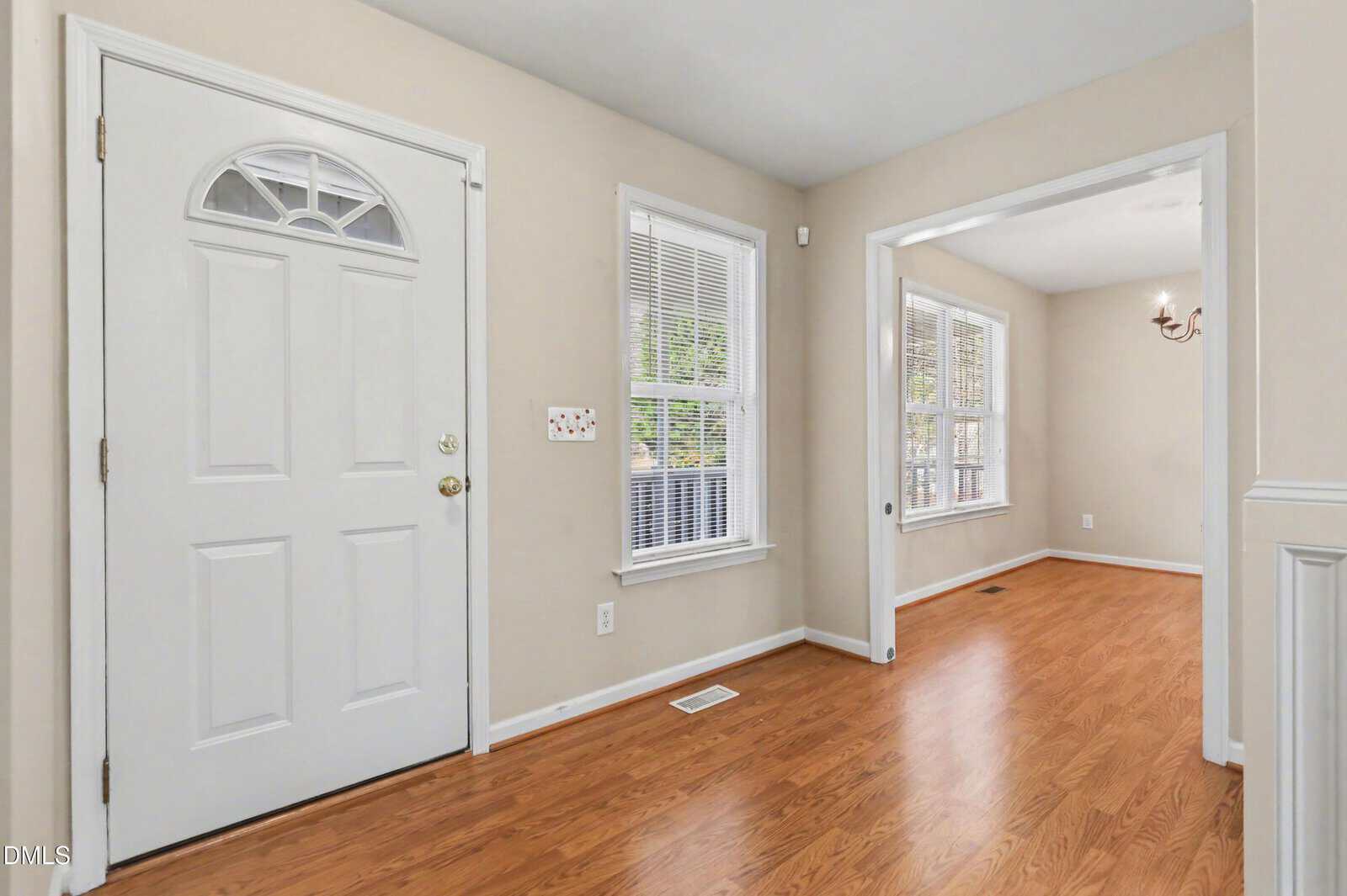 A front entry area with a white door, beige walls, wood flooring, and two windows with blinds. The space opens into an adjacent room with a small chandelier and more windows.