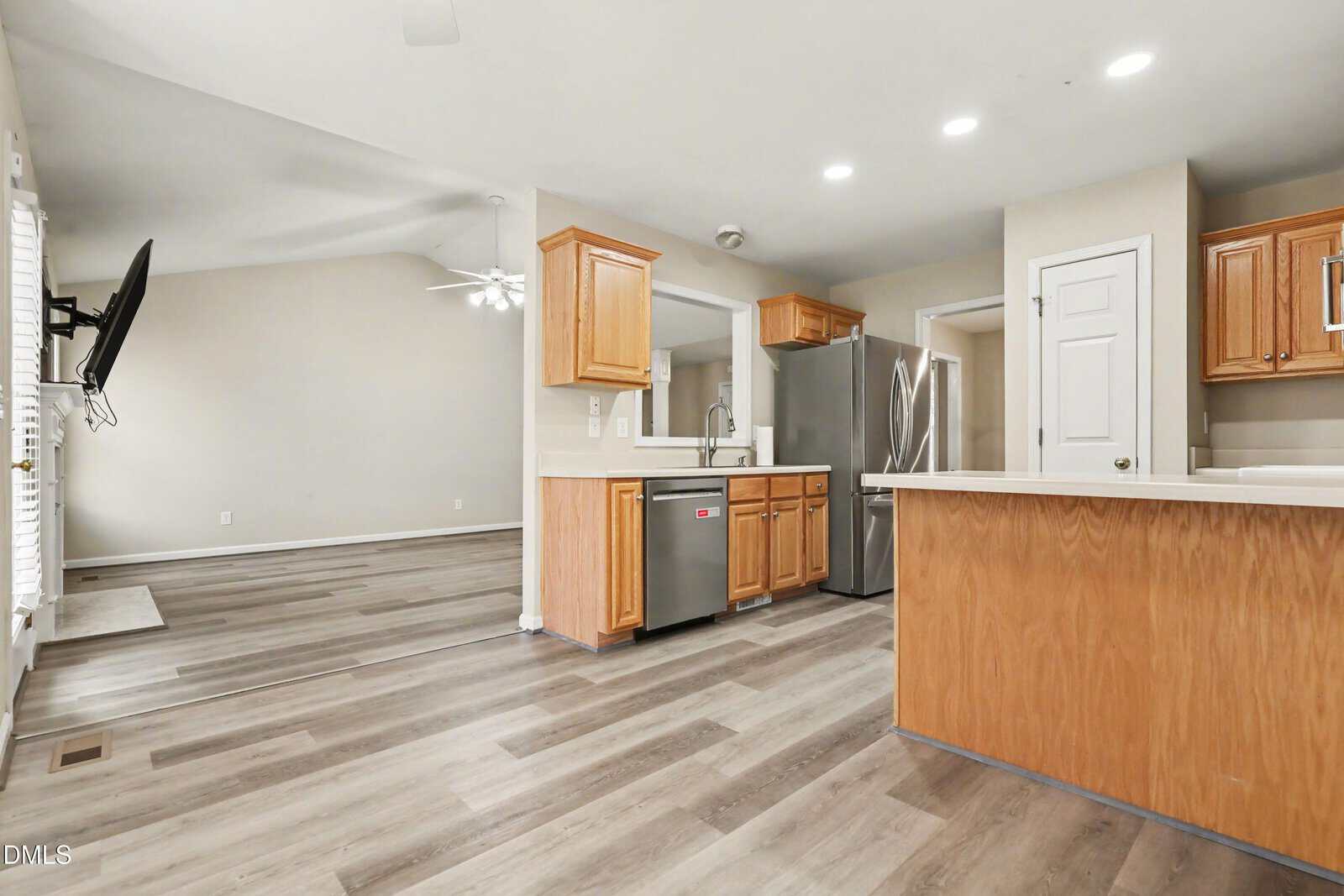 Modern kitchen with light wood cabinets, stainless steel appliances, white countertops, and vinyl plank flooring. Open layout connects kitchen to living area with ceiling fan. White walls and recessed lighting are visible throughout.