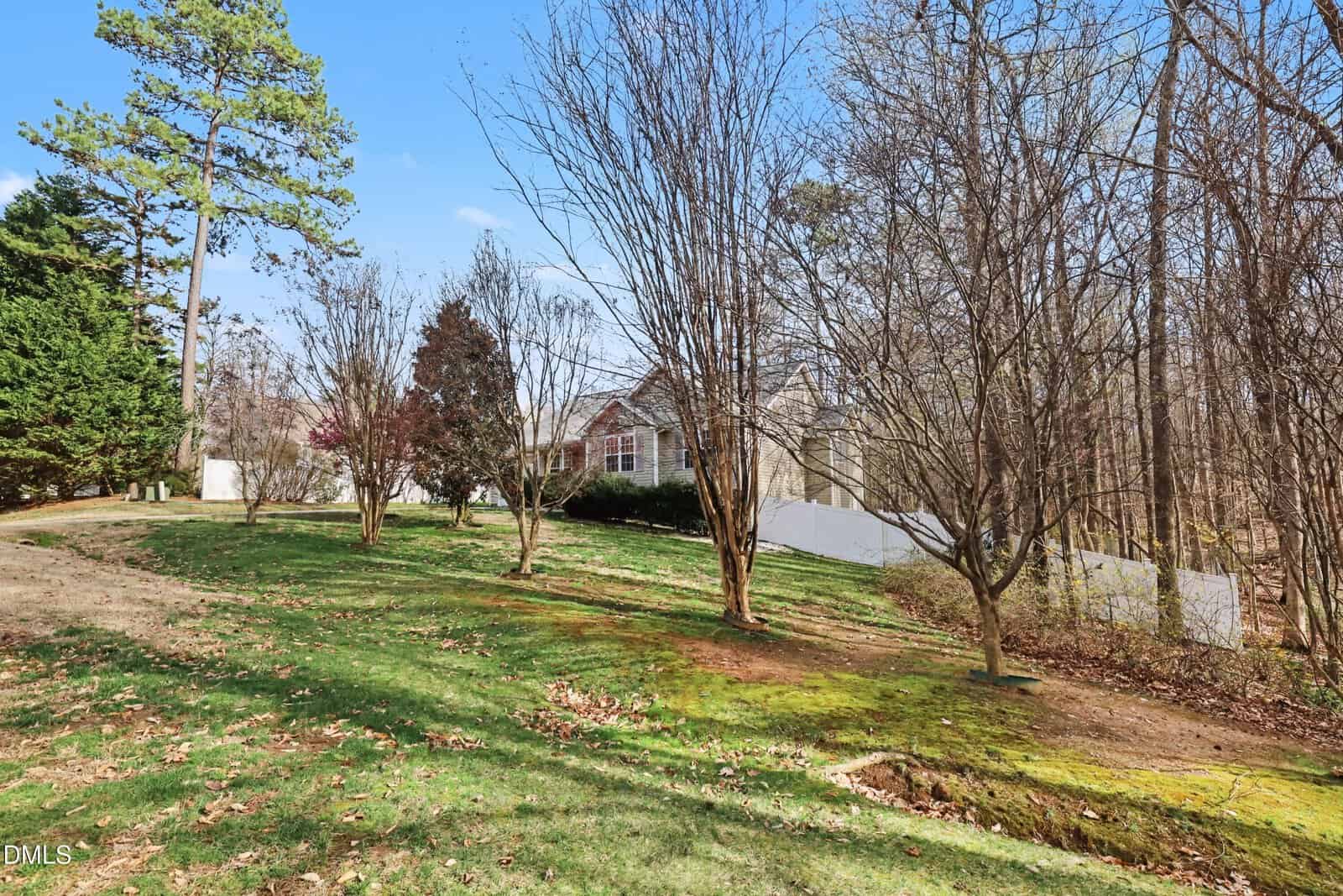 A single-story house with white siding is partially visible behind several leafless trees and shrubs. The lawn is green with patches of brown, and a white fence runs along the side of the property under a clear blue sky.
