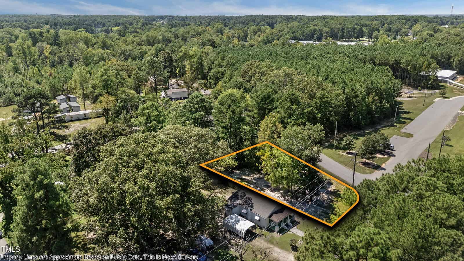Aerial view of a tree-covered residential lot outlined in orange, located at the corner of two roads in a suburban neighborhood with surrounding greenery and scattered houses.