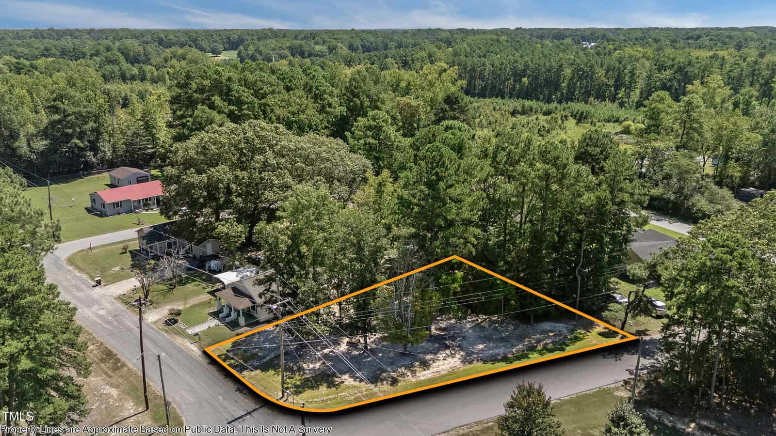 A vacant, triangular lot bordered by roads on three sides is outlined in orange among trees and houses in a suburban area, with power lines crossing overhead and forested land in the background.