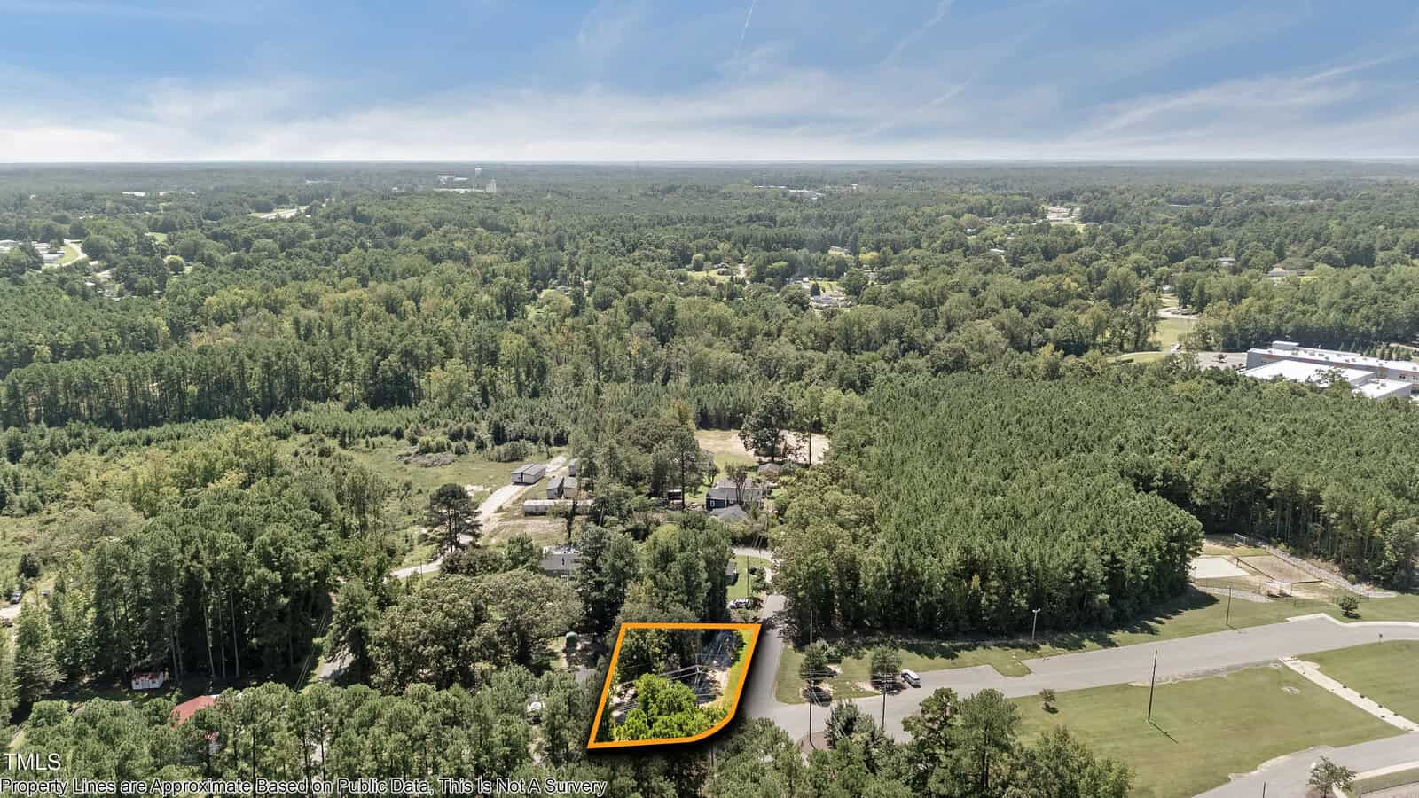 Aerial view of a wooded suburban area with a rectangular property outlined in yellow near a road; surrounding landscape features dense trees, scattered houses, and clear skies.