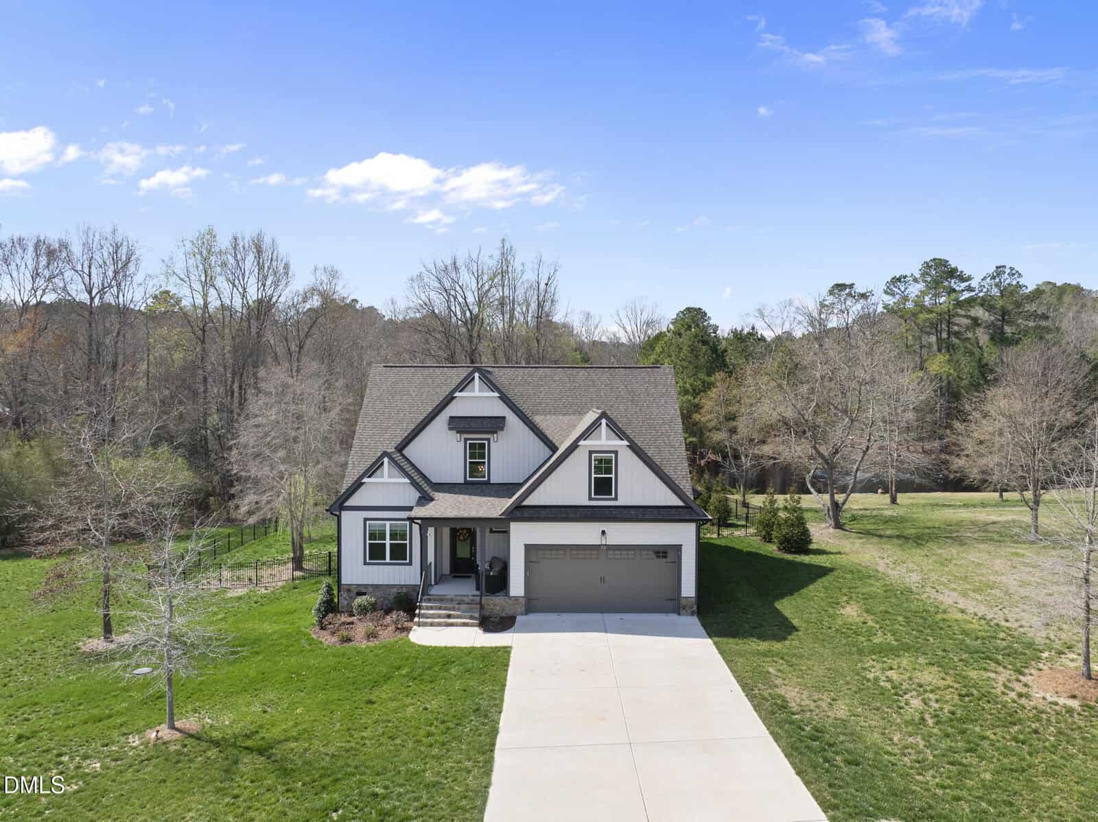 A two-story house with a gabled roof, covered front porch, and attached two-car garage, set on a large grassy lot with trees and a concrete driveway, under a partly cloudy sky.