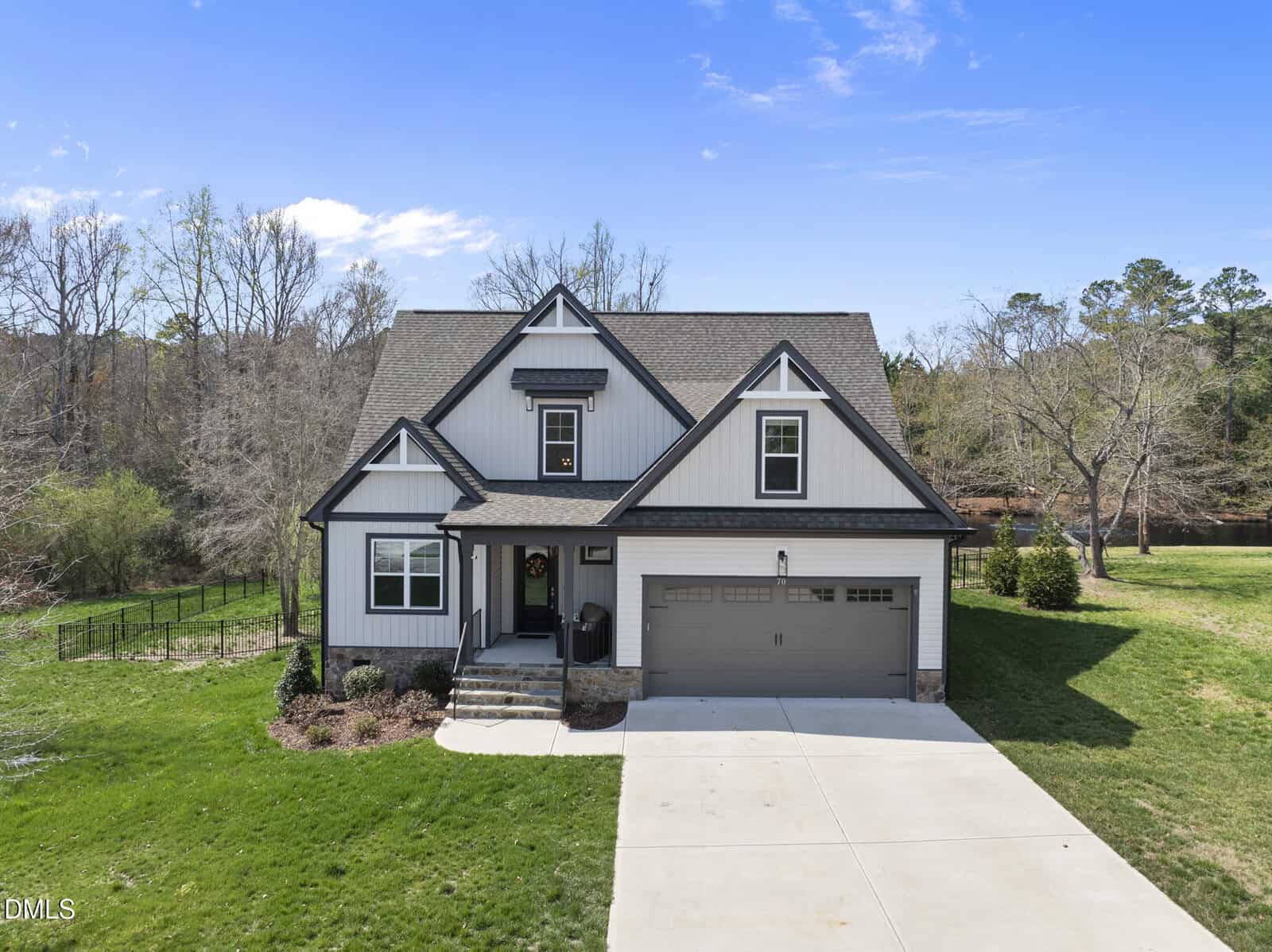 A modern two-story house with gray siding, dark trim, and a two-car garage sits on a well-kept lawn with a concrete driveway; trees and shrubs surround the property under a clear sky.
