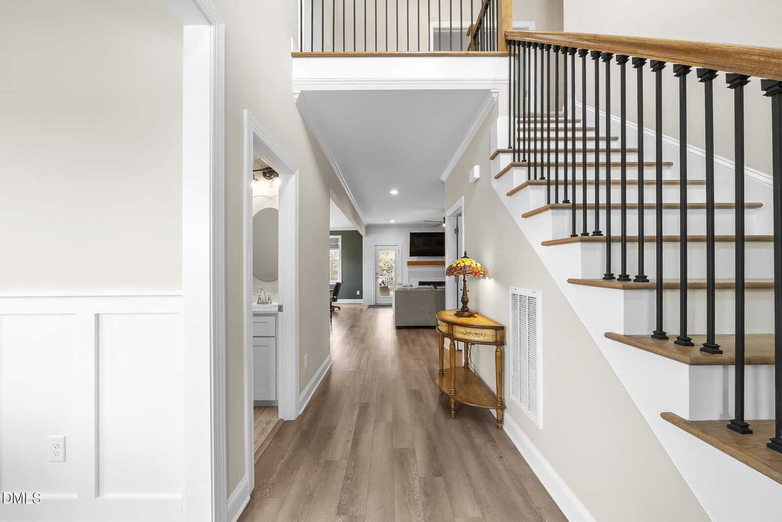 A hallway with light wood flooring, a staircase with black railings, a small wooden table with a flower vase, and a view into a living room with a TV and windows. A bathroom is visible on the left.