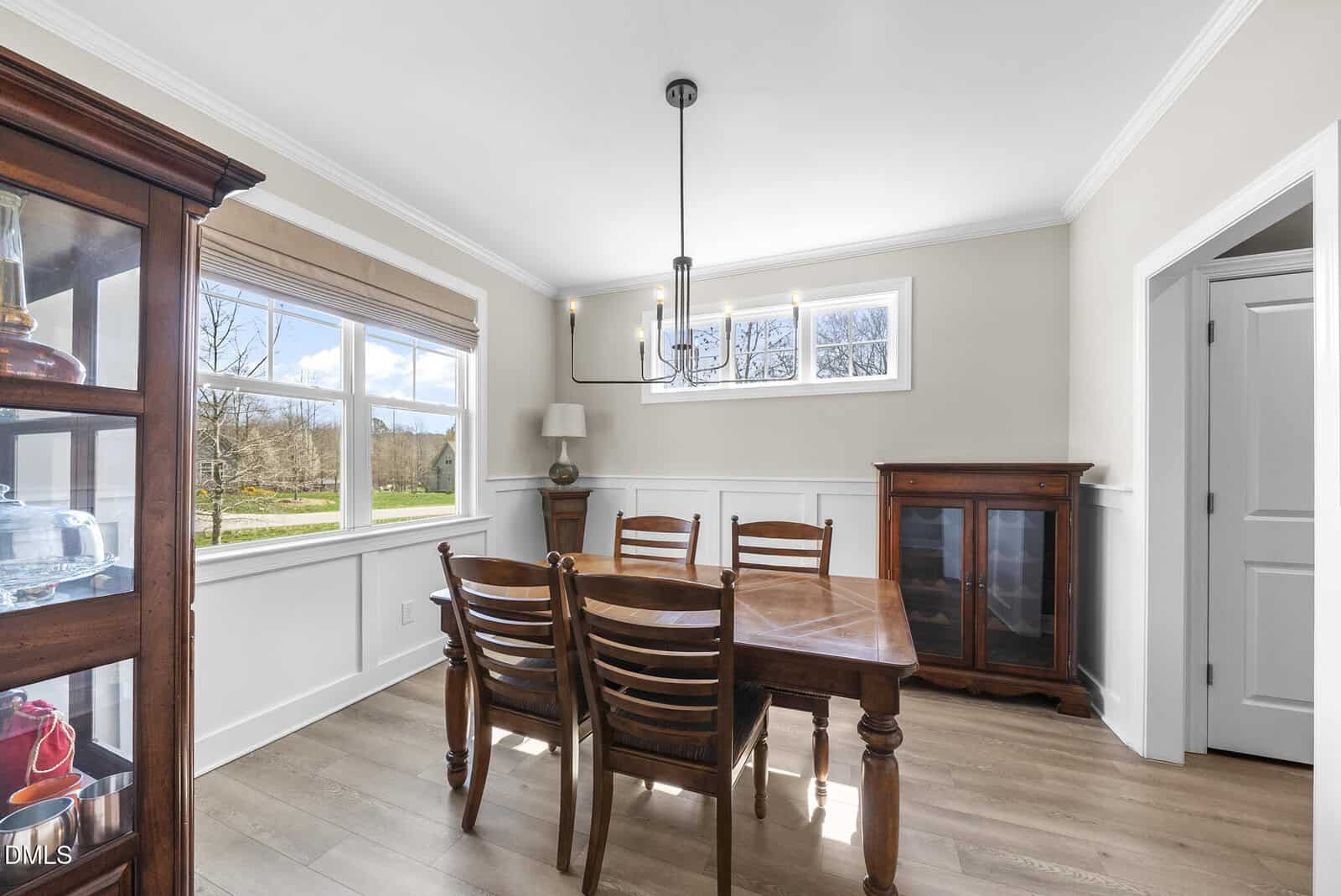 A dining room with a wooden table and four chairs, glass-fronted display cabinets, wood flooring, large windows, and a modern light fixture hanging from the ceiling.