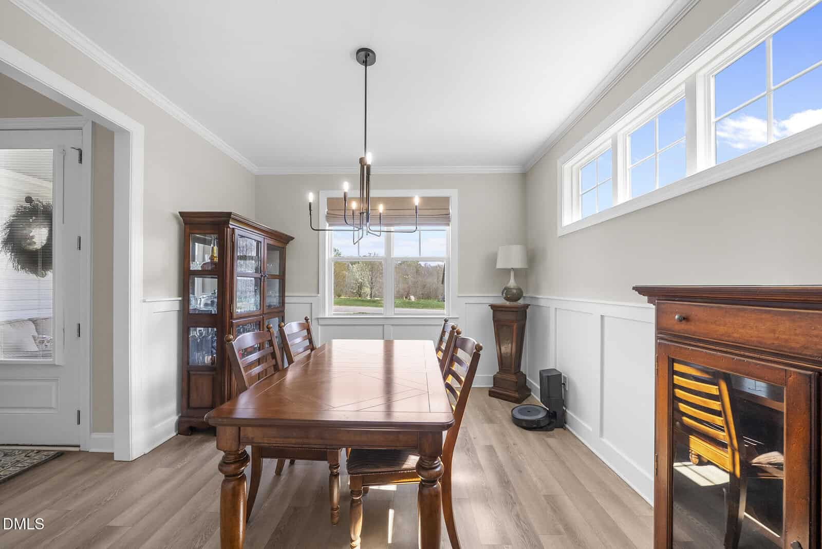 Bright dining room with wooden table and chairs, glass cabinet, chandelier, and large windows letting in sunlight. Wood floors and neutral walls create a warm, inviting atmosphere.